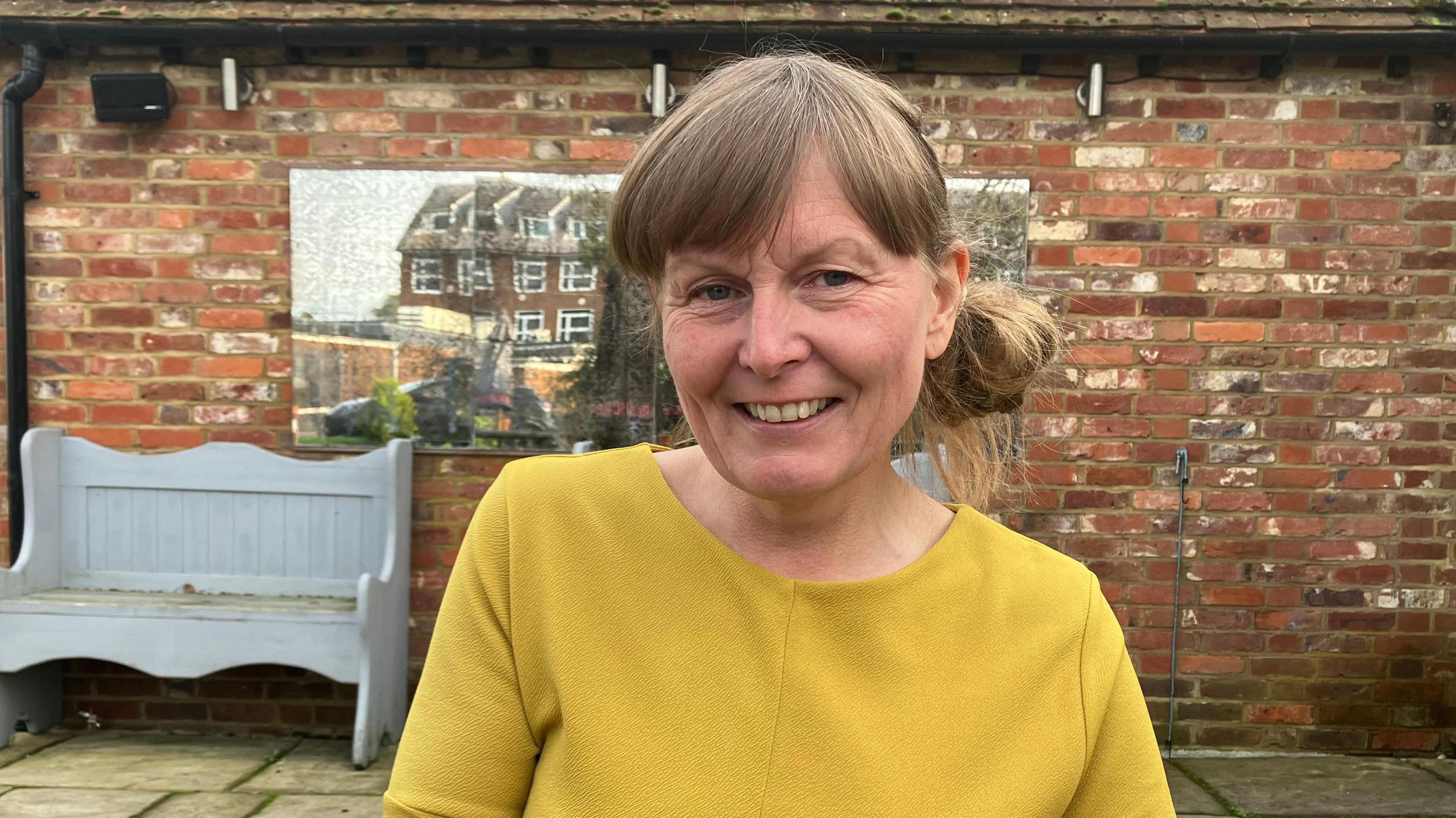 Catherine Bunting with light brown hair, tied back, with a fringe. She is wearing a mustard yellow tunic and is sat down in a pub beer garden.