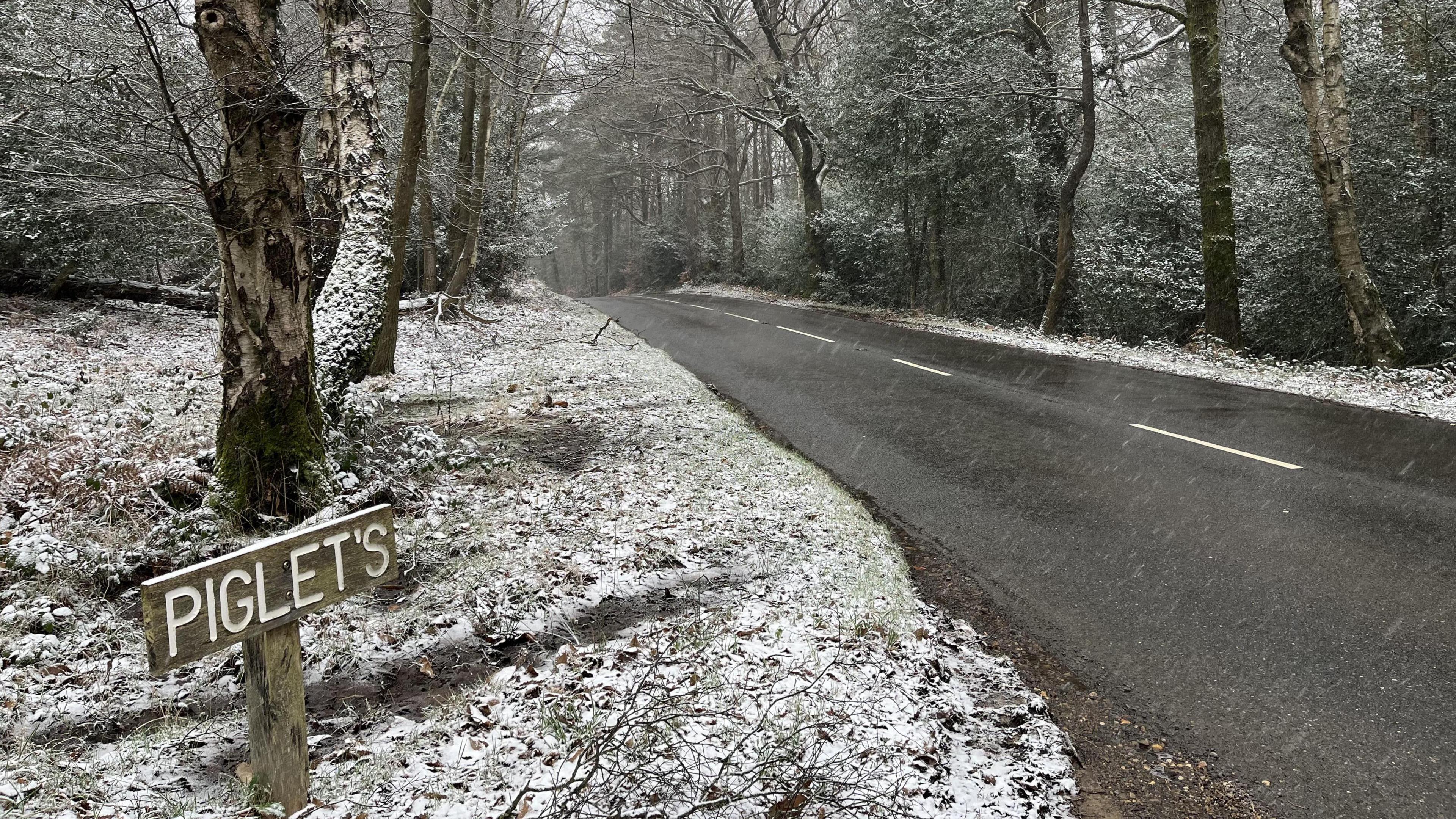 A road with a snowy forest either side of it. There is a wooden sign in the bottom left which says "PIGLET'S".