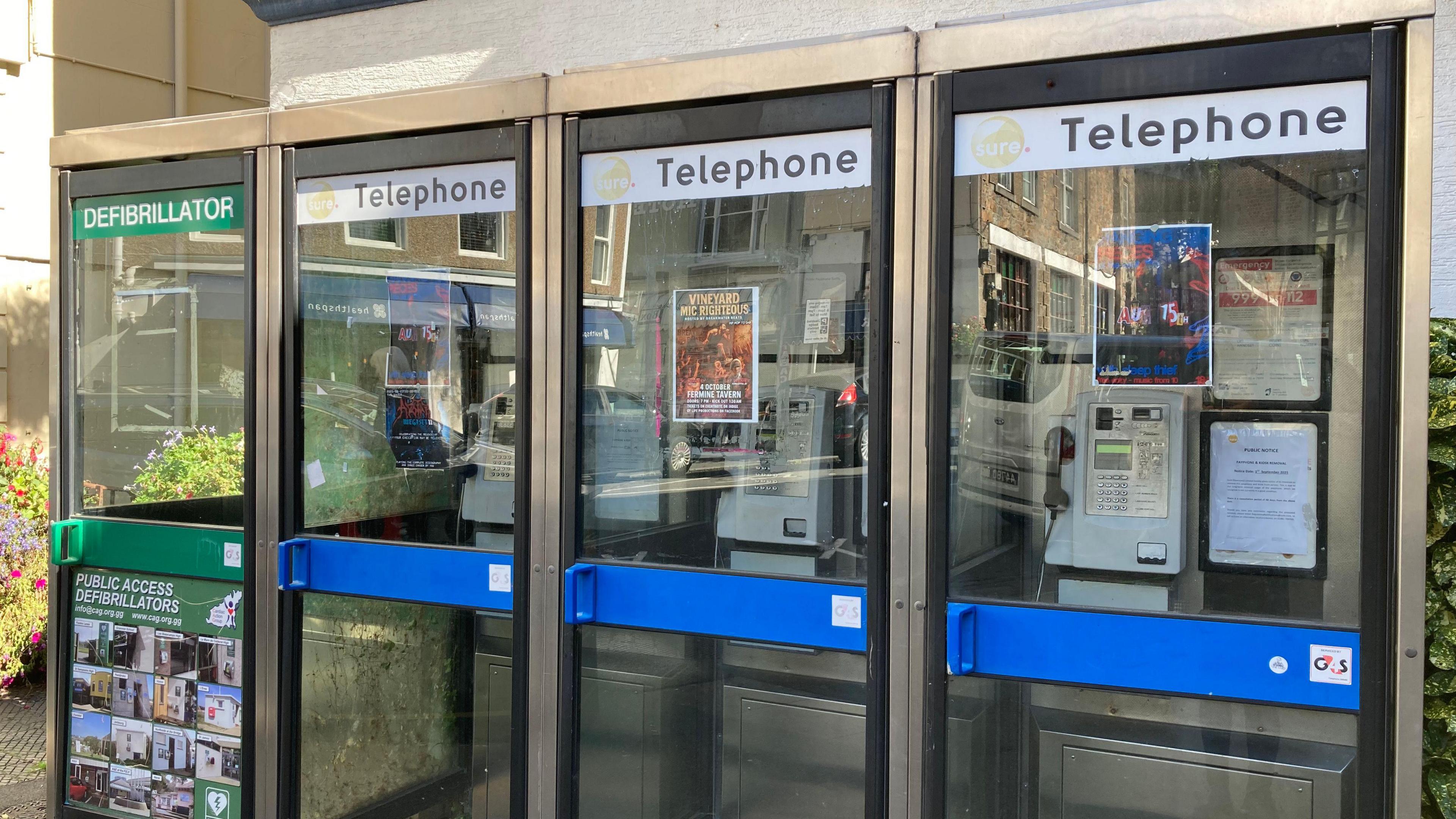 Exterior of a row of telephone boxes. The payphones are inside a glass structure. The handle to open the door is blue.