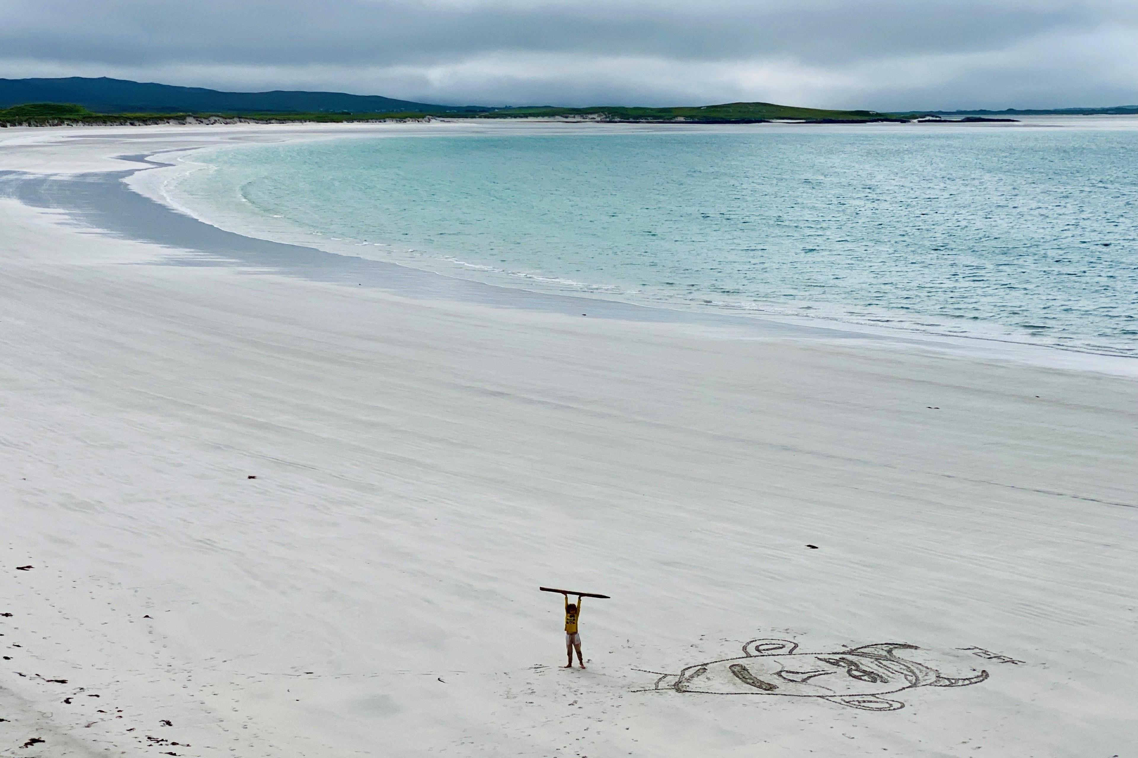 A boy stands on a huge, empty, white beach holding up a stick he has used to draw a face on the sand.