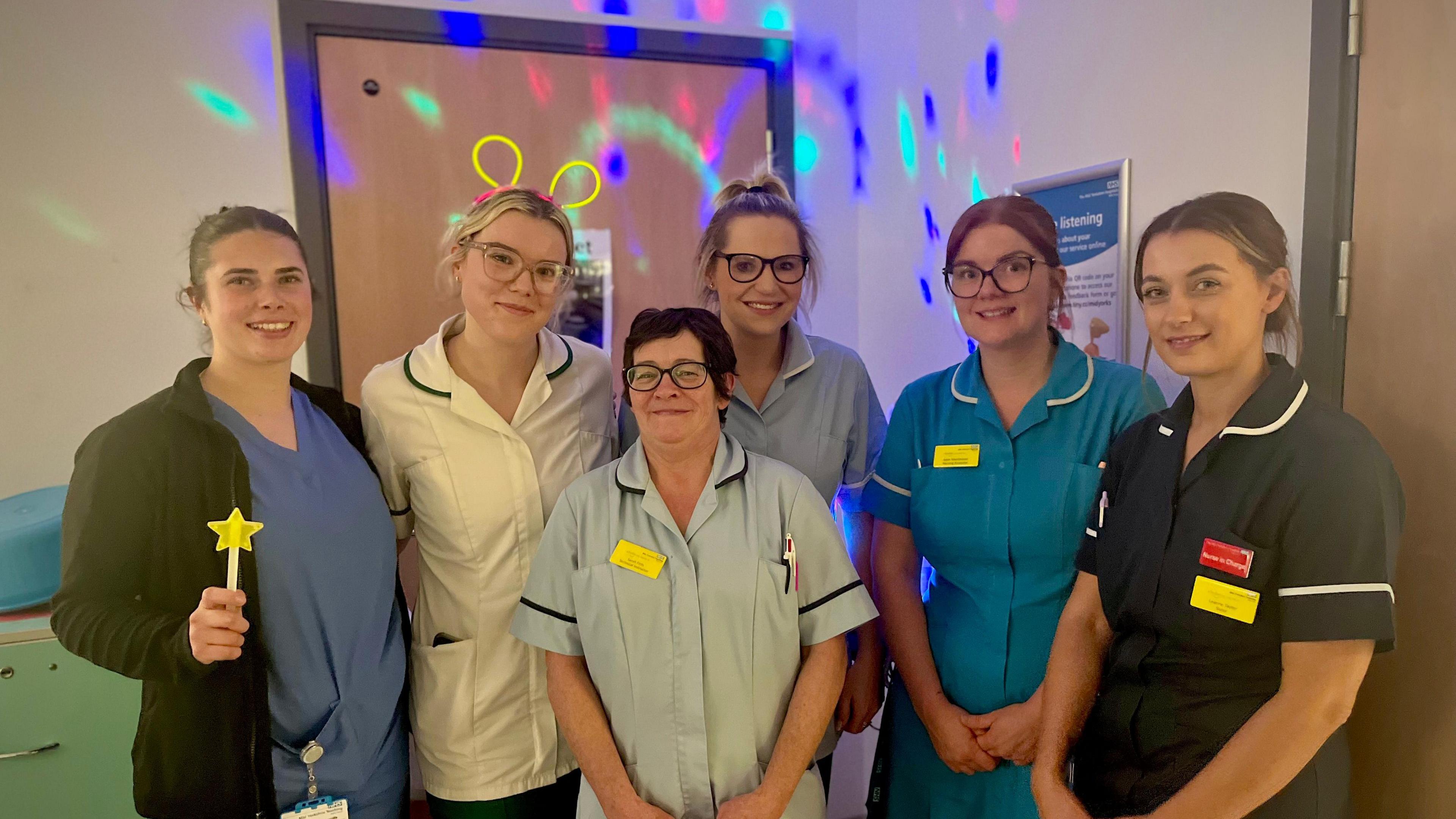 Six women in nurses' scrubs stand in a ward. Some are wearing glow sticks. There are soft bright projected lights behind them.