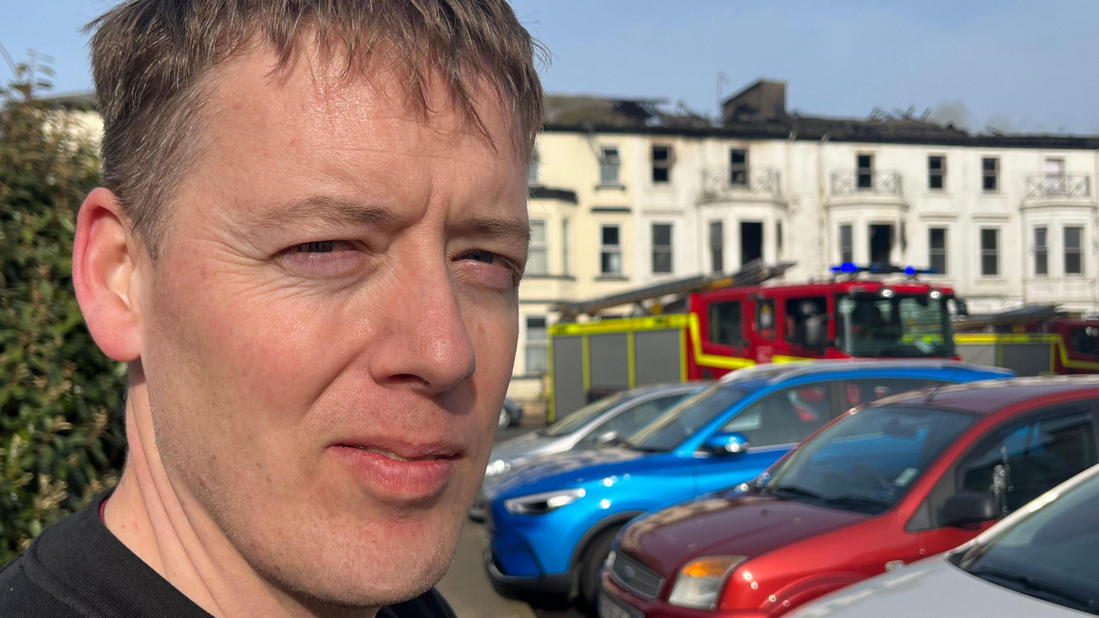 A man with fair hire wearing a black t-shirt faces the camera with a fire engine and a fire-damaged building in the background.