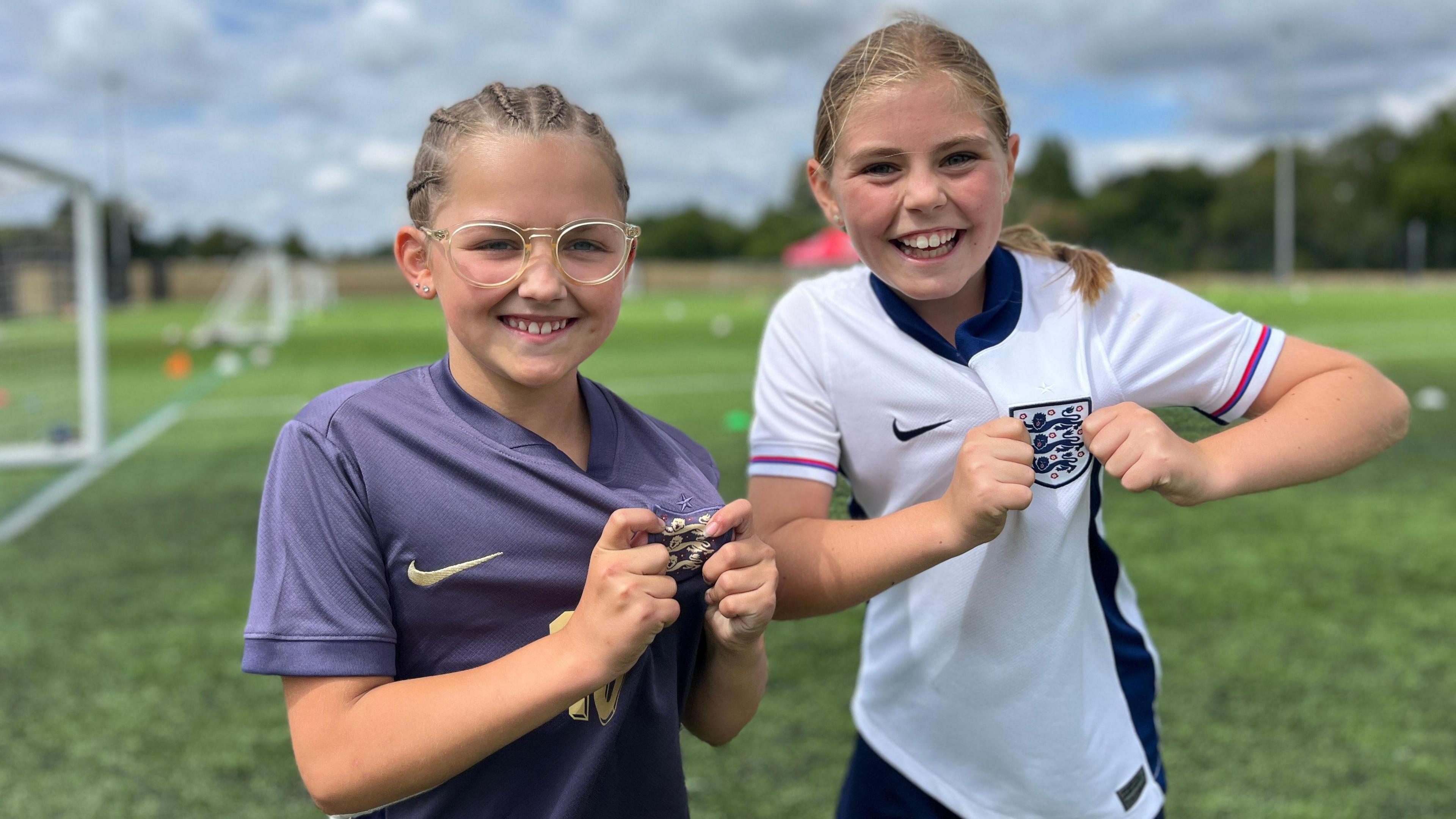 Two young girls, both of them wearing England football tops, one grey and one white. They are both holding the three lions crest on their tops as they smile for the camera.