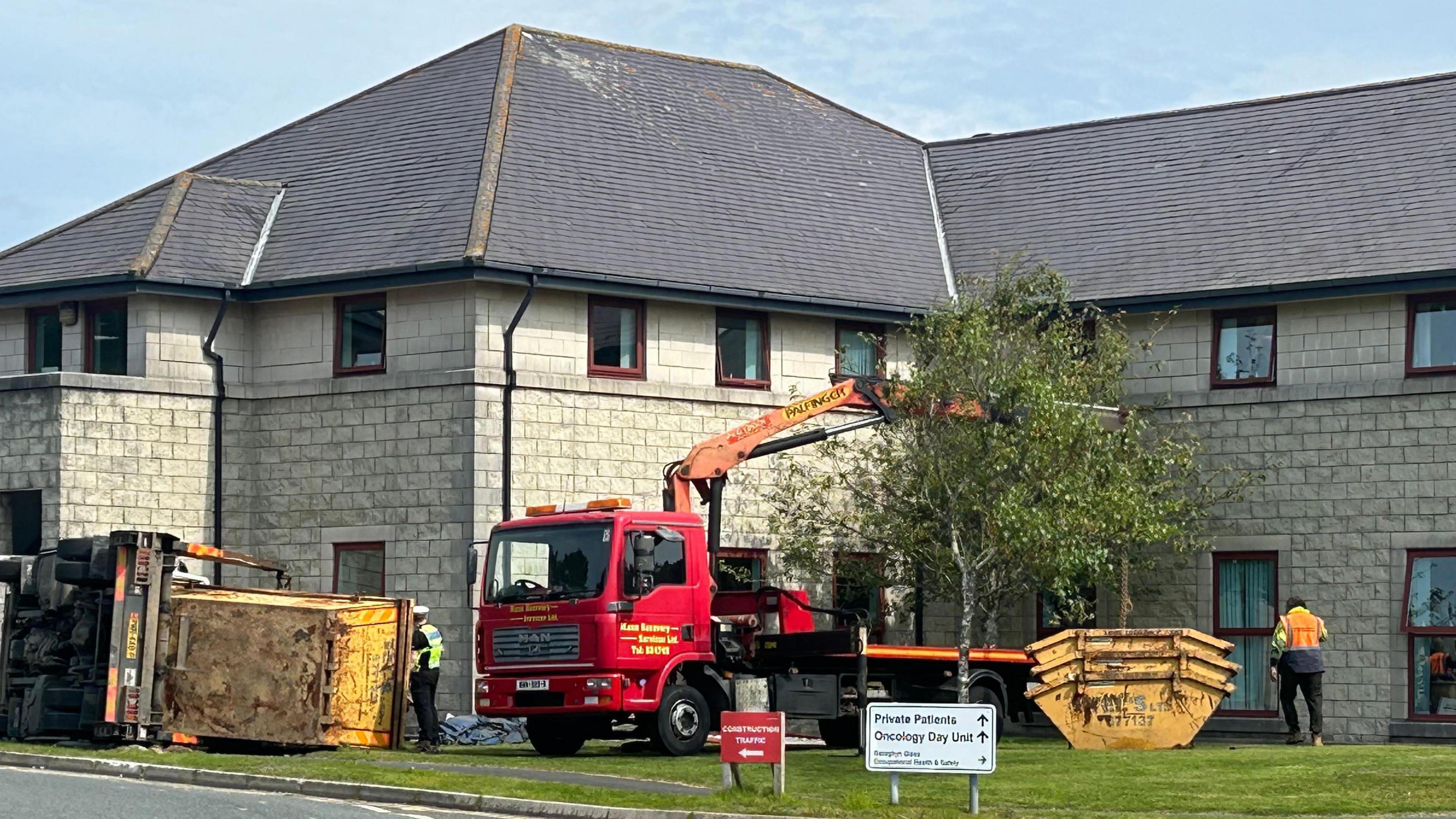 A building in the background, you can see a red truck with a crane on a field in front, a skip truck has tipped over on its side.