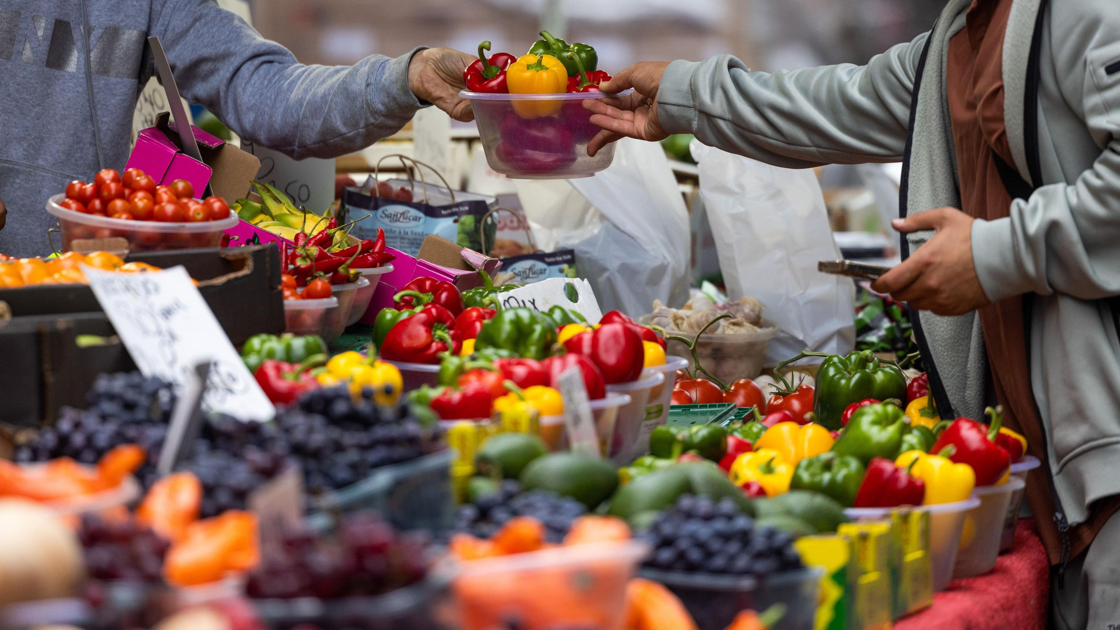 A trader hands over a punnet of vegetables to a customer on a market stall, filled with fruit and vegetables.