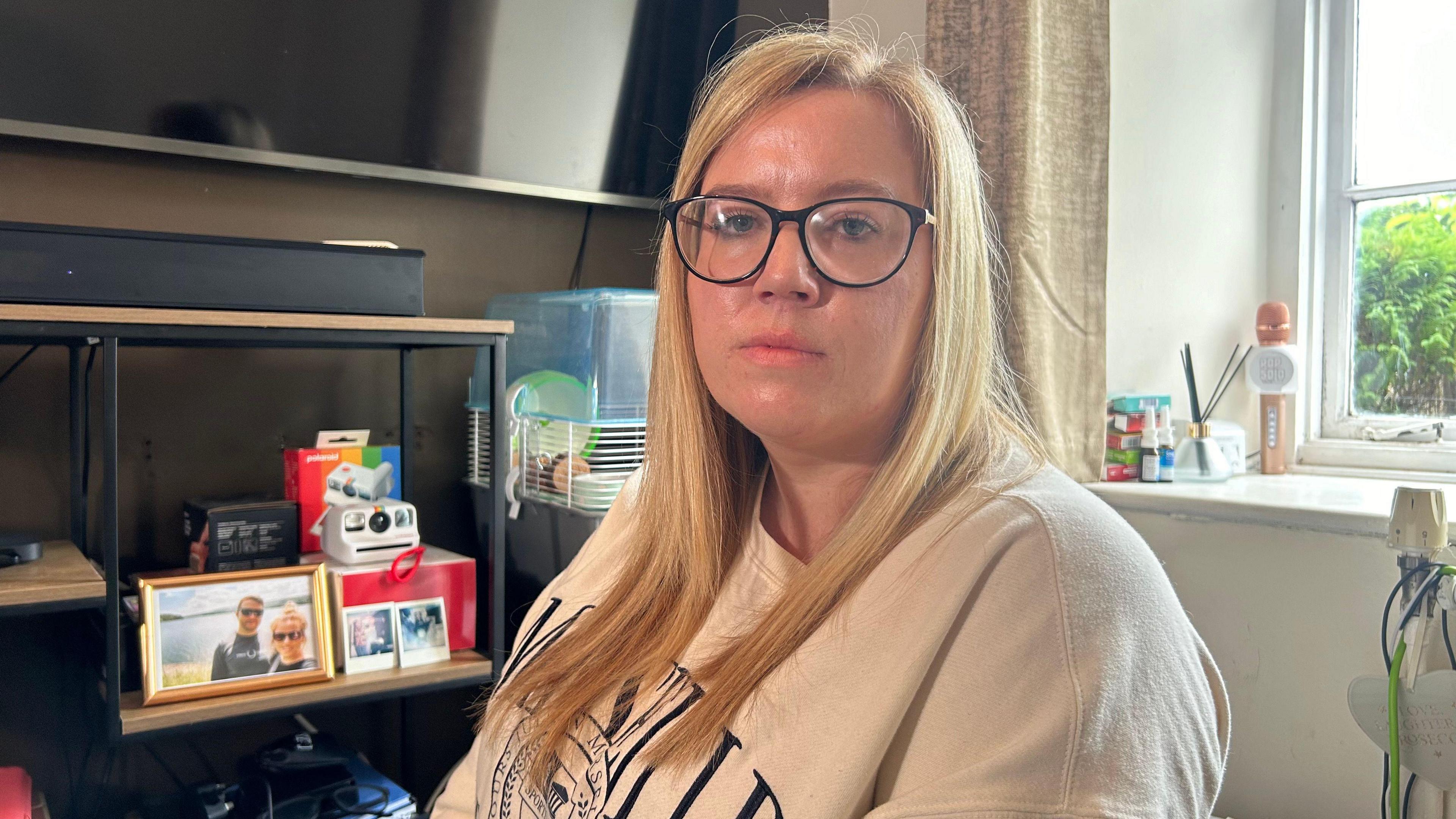 A woman who has blonde hair and glasses, sitting in her living room with a TV and photos in the background.