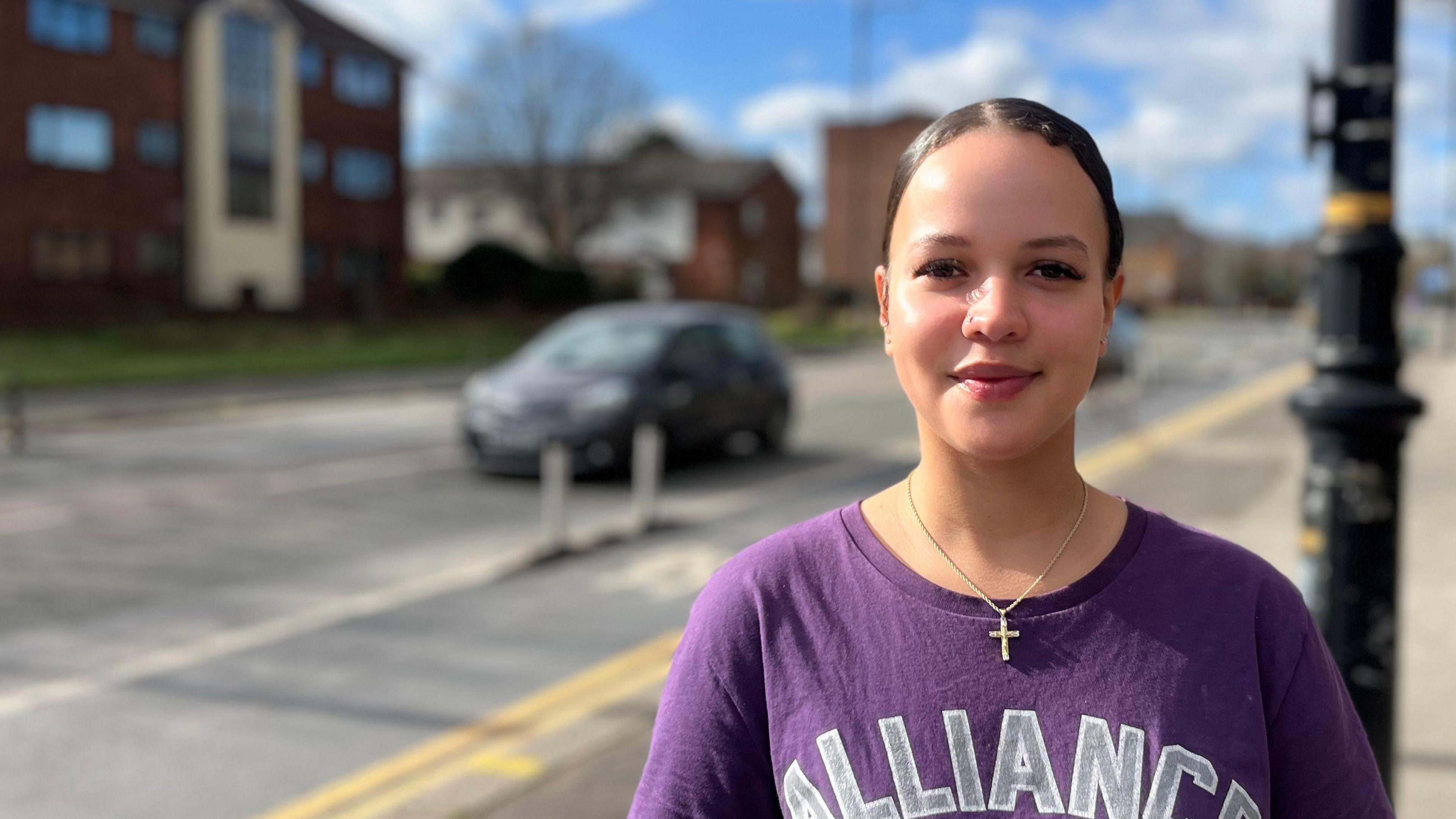 Amelia with brown hair tied back wearing a purple t-shirt with a silver cross in a street near Old Trafford