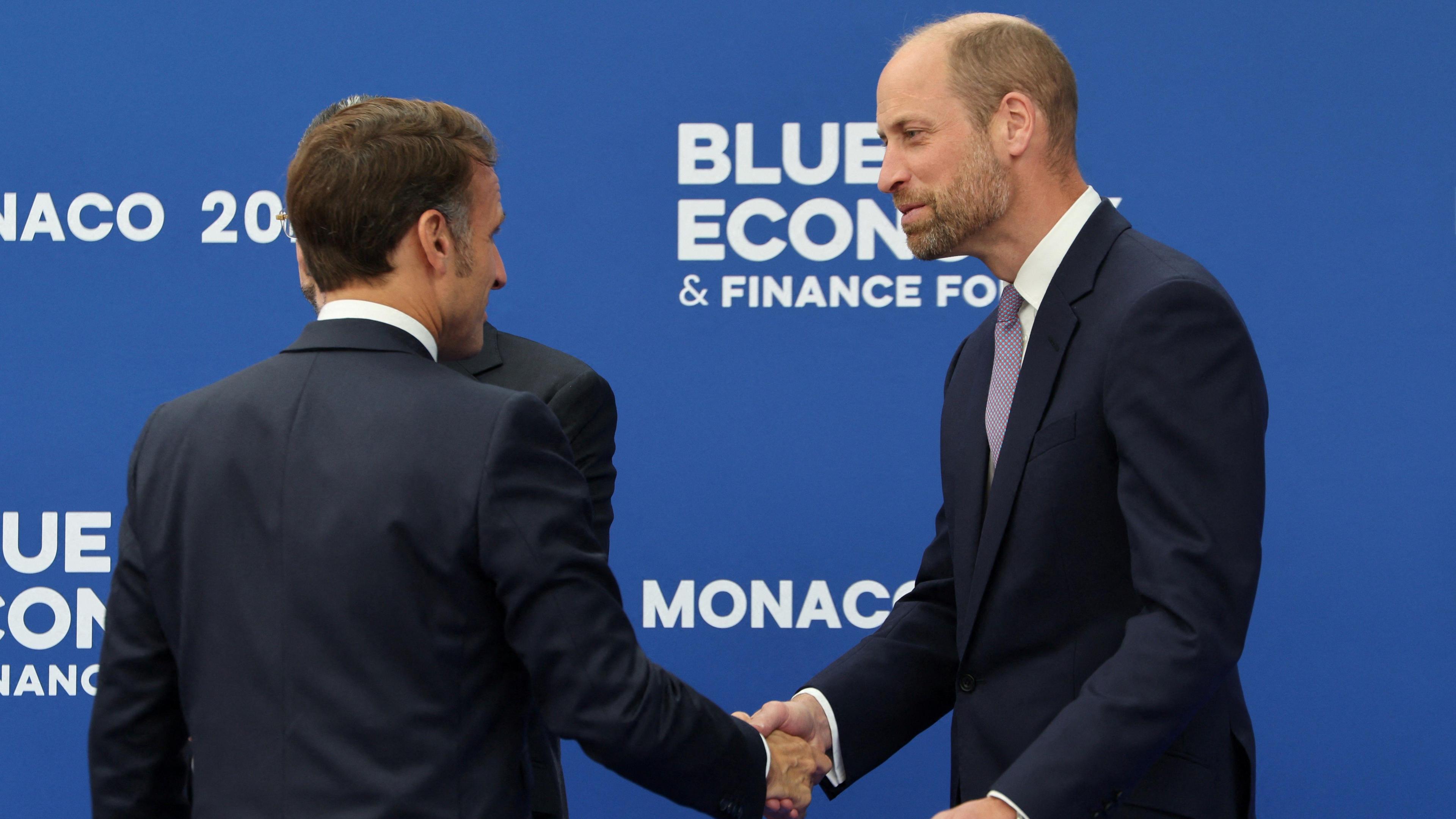 French President Emmanuel Macron shaking hands with Prince William in front of a blue background