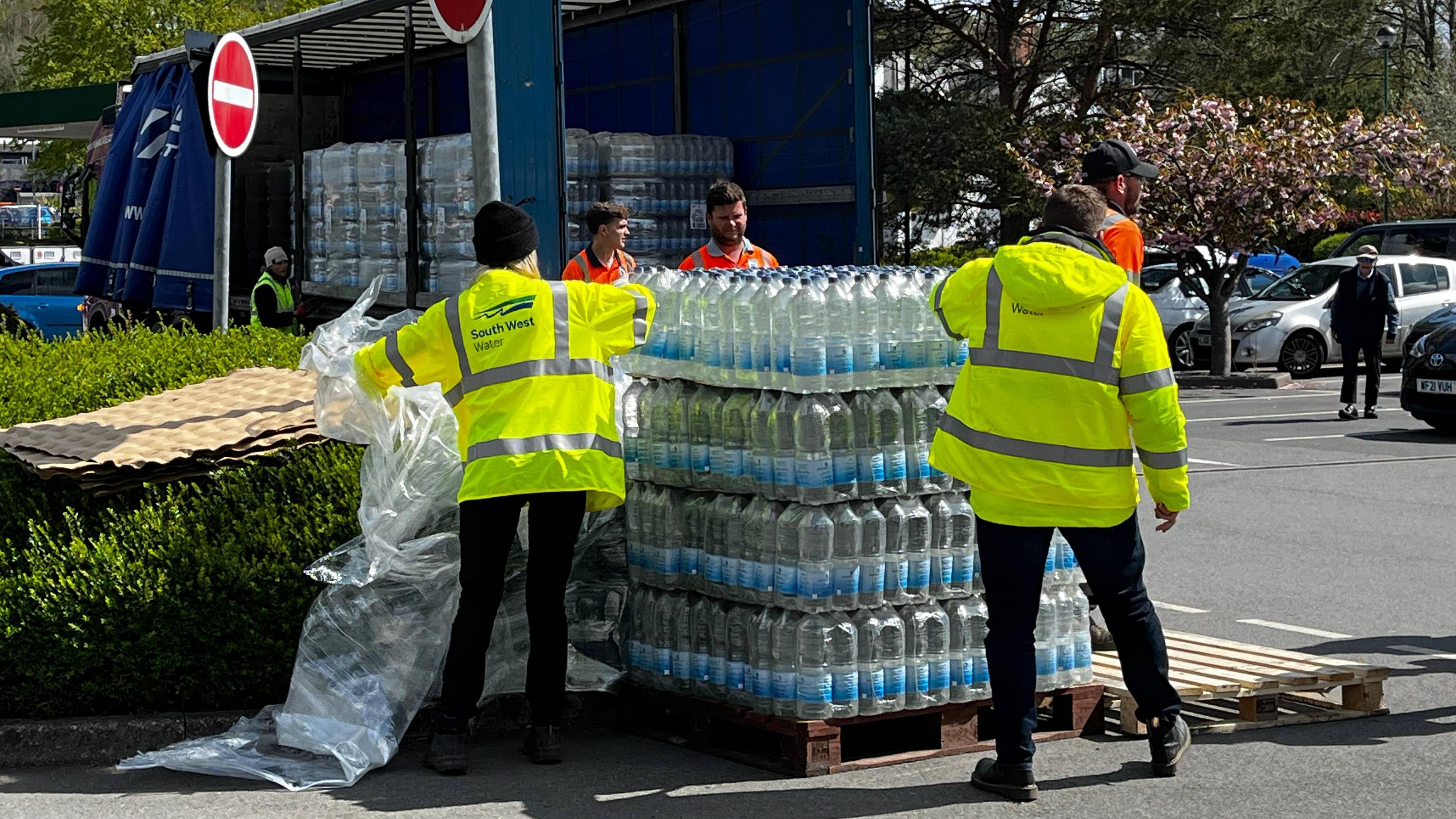 Bottled water stacked on a wooden pallet in front of a blue lorry. The lorry is open, where there is more large bottles of water. There are two people wearing yellow hi-vi's, one with the words 'South West Water' on the back and three others wearing orange hi-vi's. They are stood in the Morrison's carpark in Tavistock.