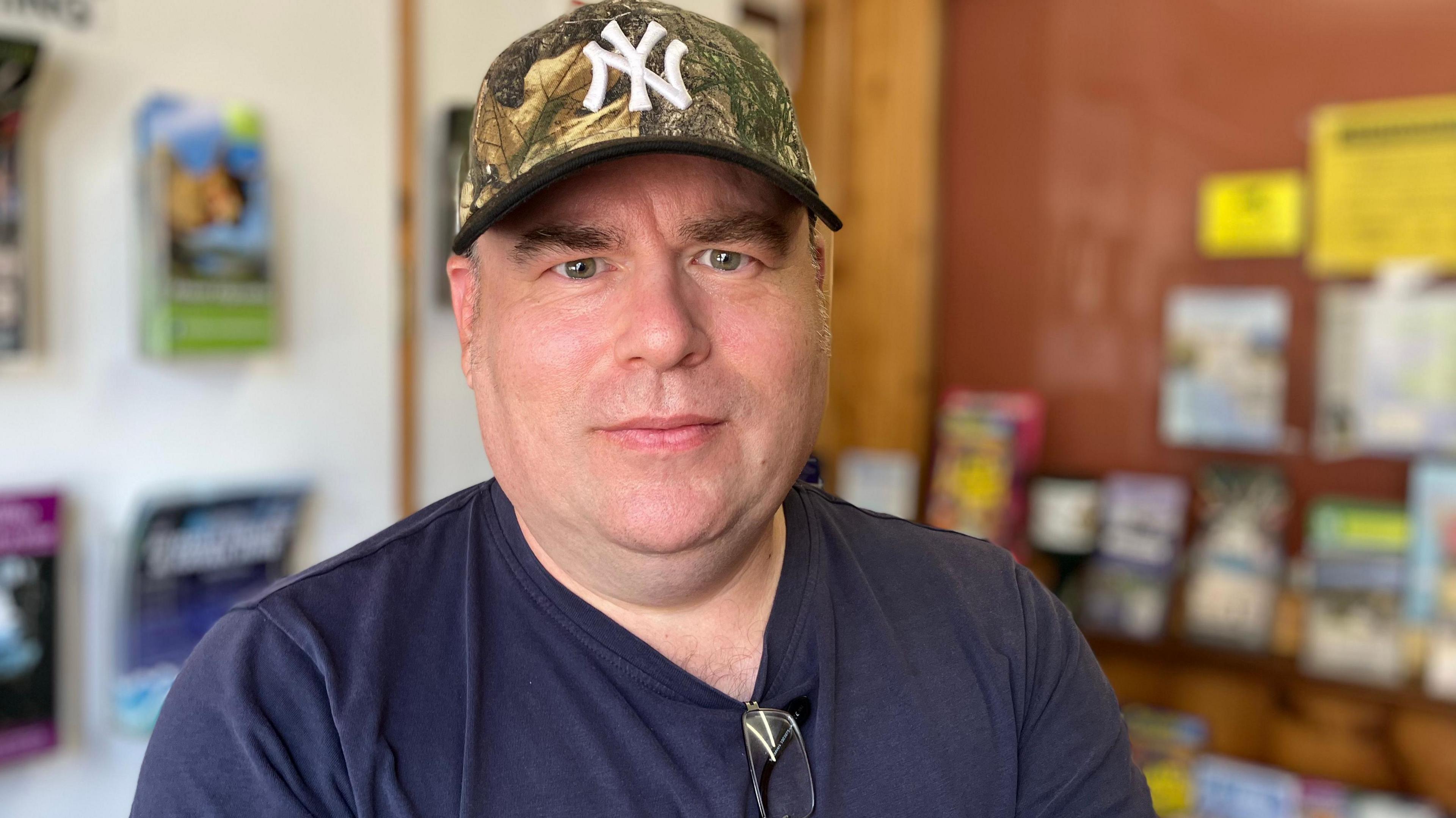 A man in a dark blue top and khaki baseball cap looks straight at the camera.
Tourist leaflets can be seen in holders in soft focus behind him.