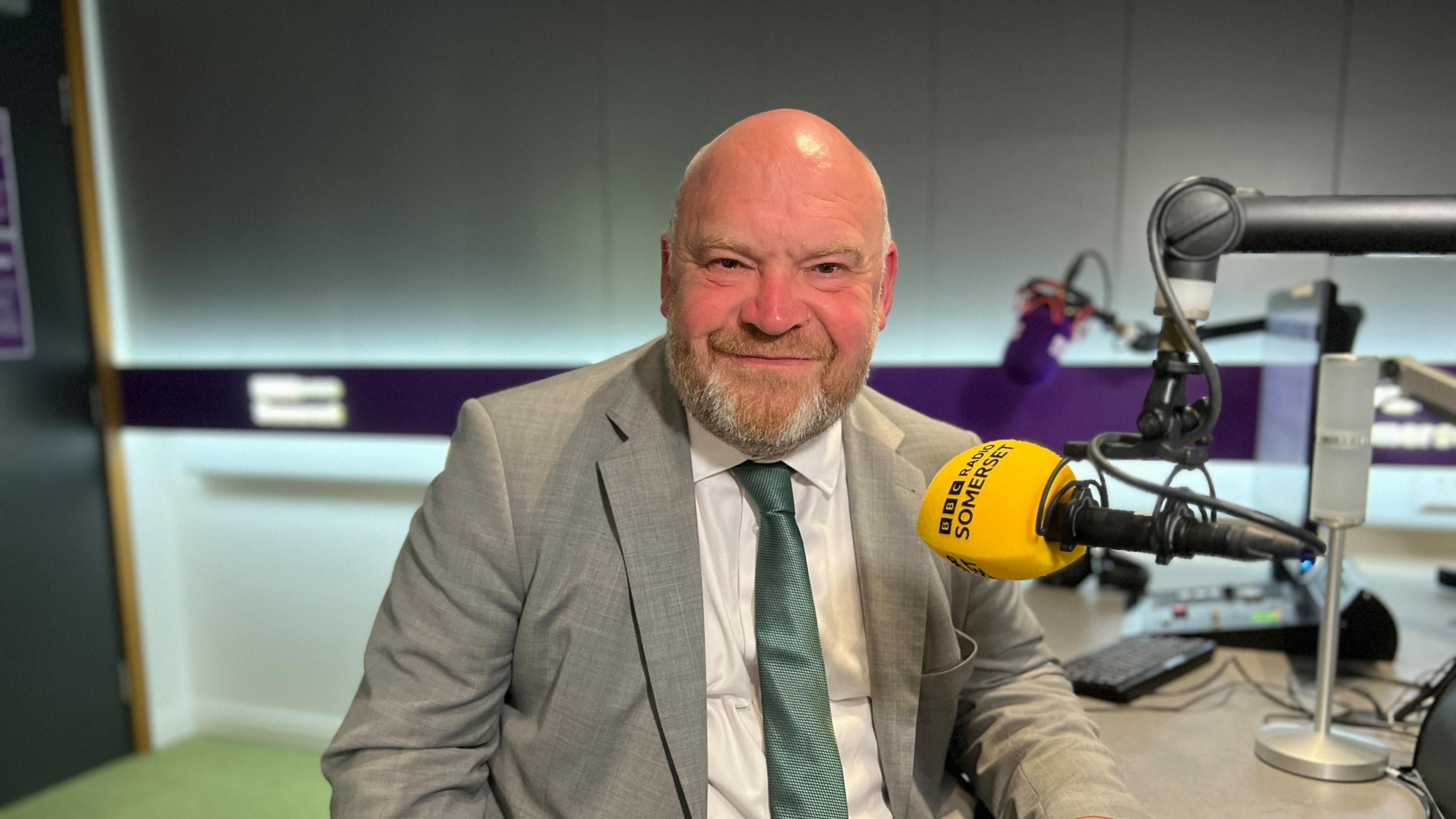 Bill Revans in a tie and suit jacket sitting behind a yellow BBC Radio Somerset microphone