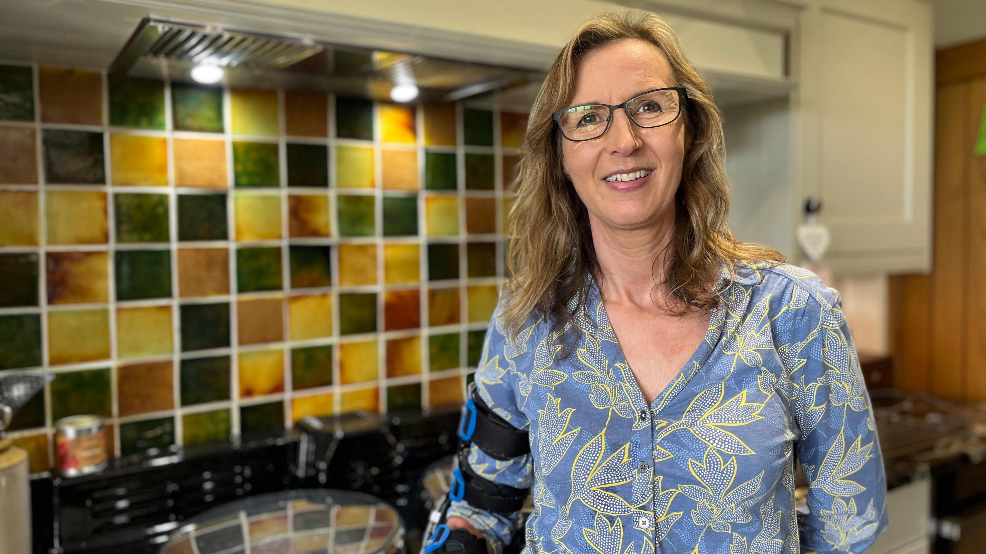 Jayne Jones has brown/blonde hair and is wearing a blue shirt. She is standing in a kitchen.