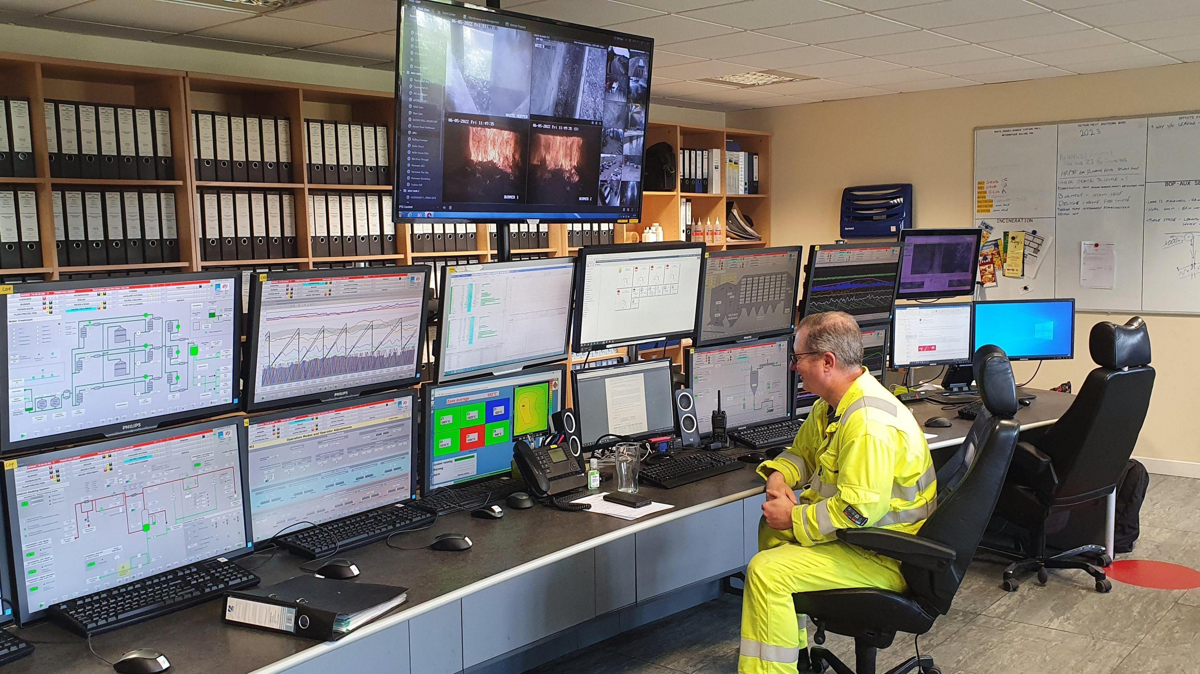 A man in a high visibility outfit is sat in front of screens monitoring the incinerator. There are 16 screens in total in front of him. He is sat in a black chair.