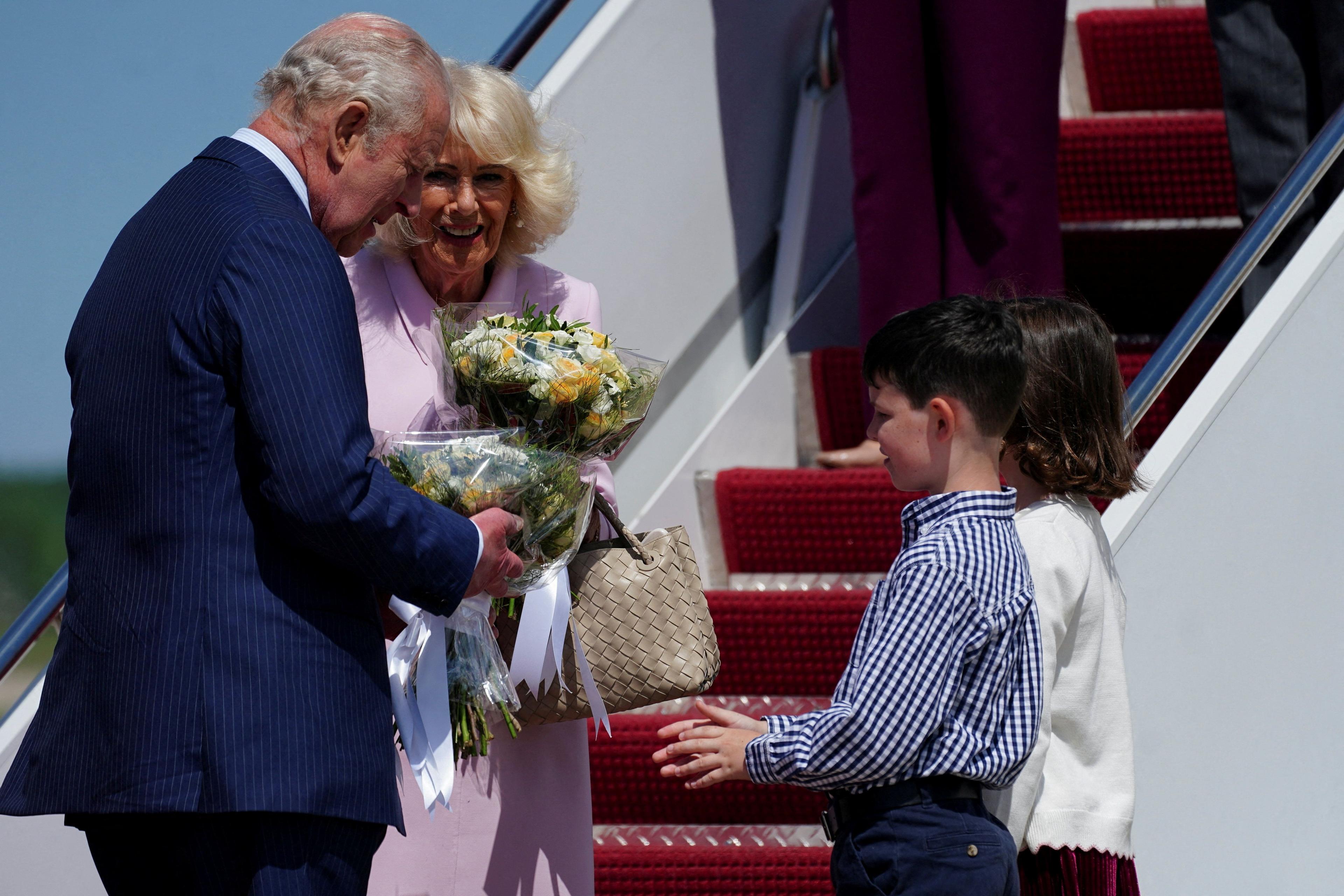 King and Queen standing at the bottom of airplane steps holding bouquets handed to them by two children. 