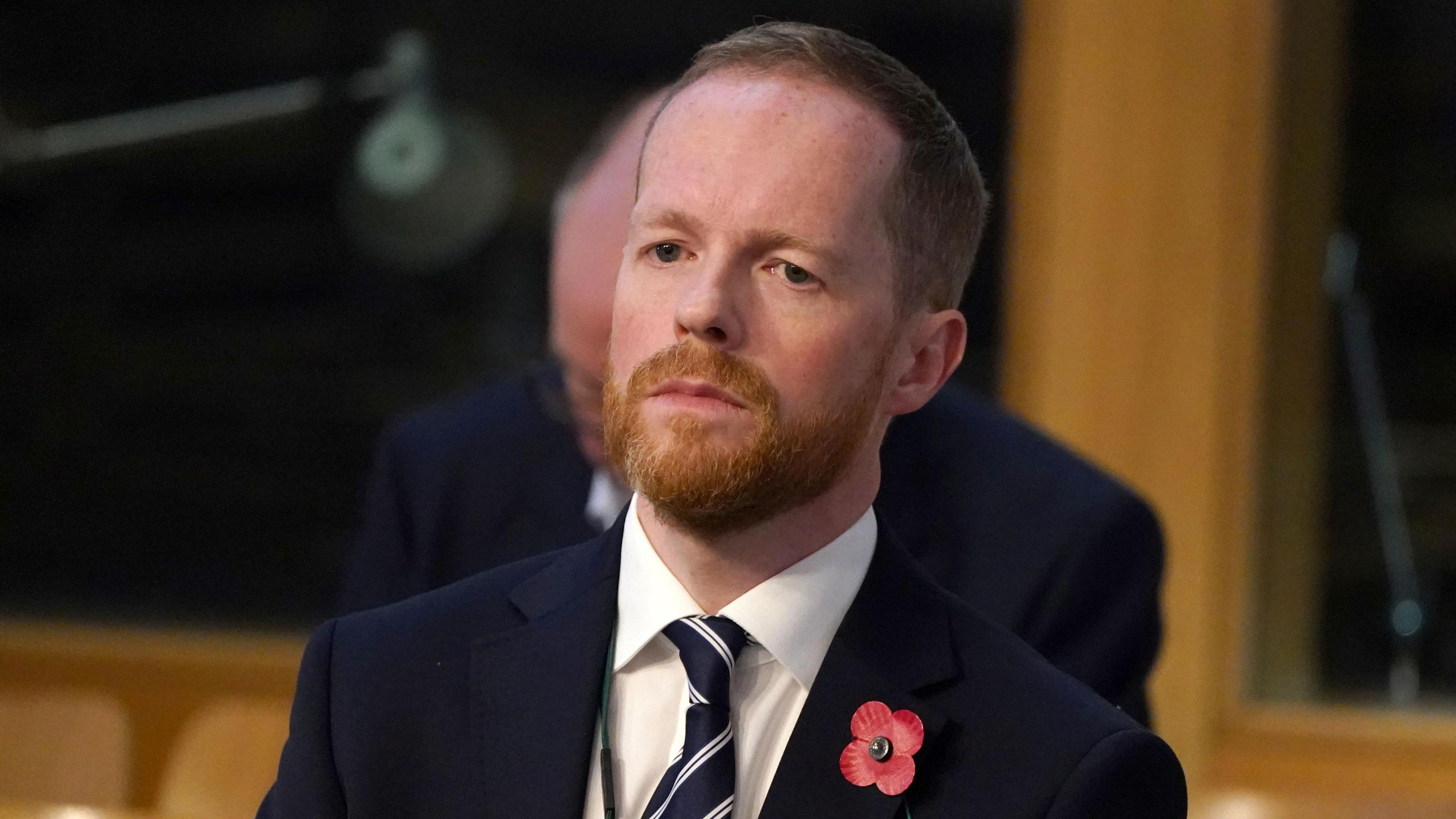 A man wearing a dark suit and a poppy, with a ginger beard and short, close cropped hair. He has a serious expression on his face, and appears to be standing in a meeting room with wood panelling.