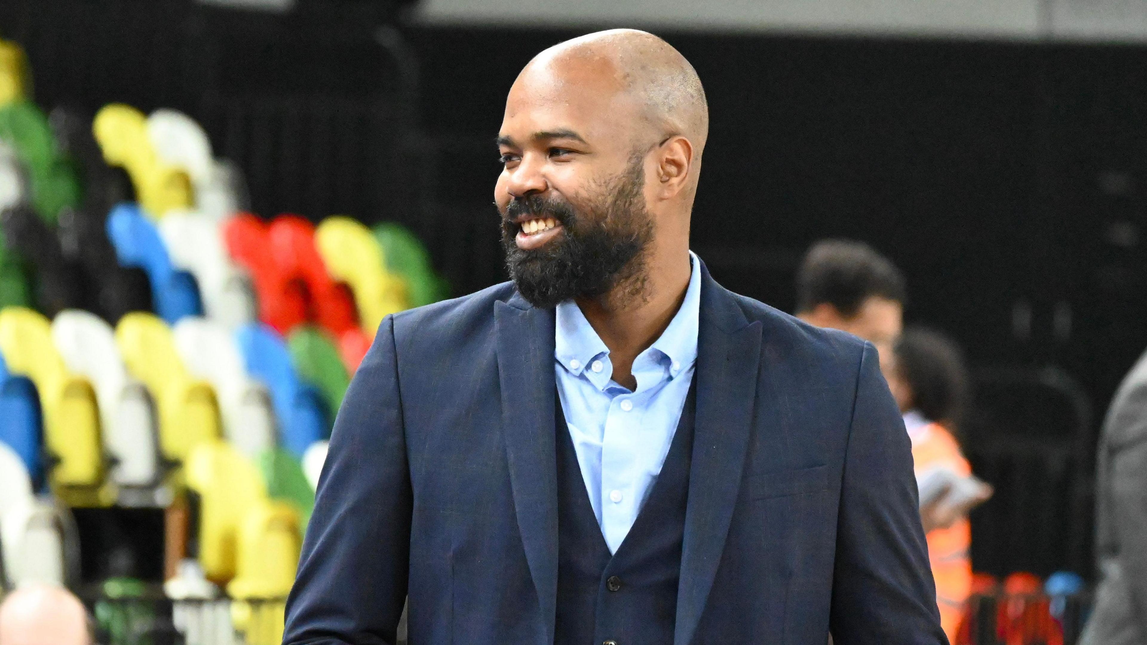 A bald man with a beard wearing a three-piece navy suit. There are multi-coloured spectator seats behind him.