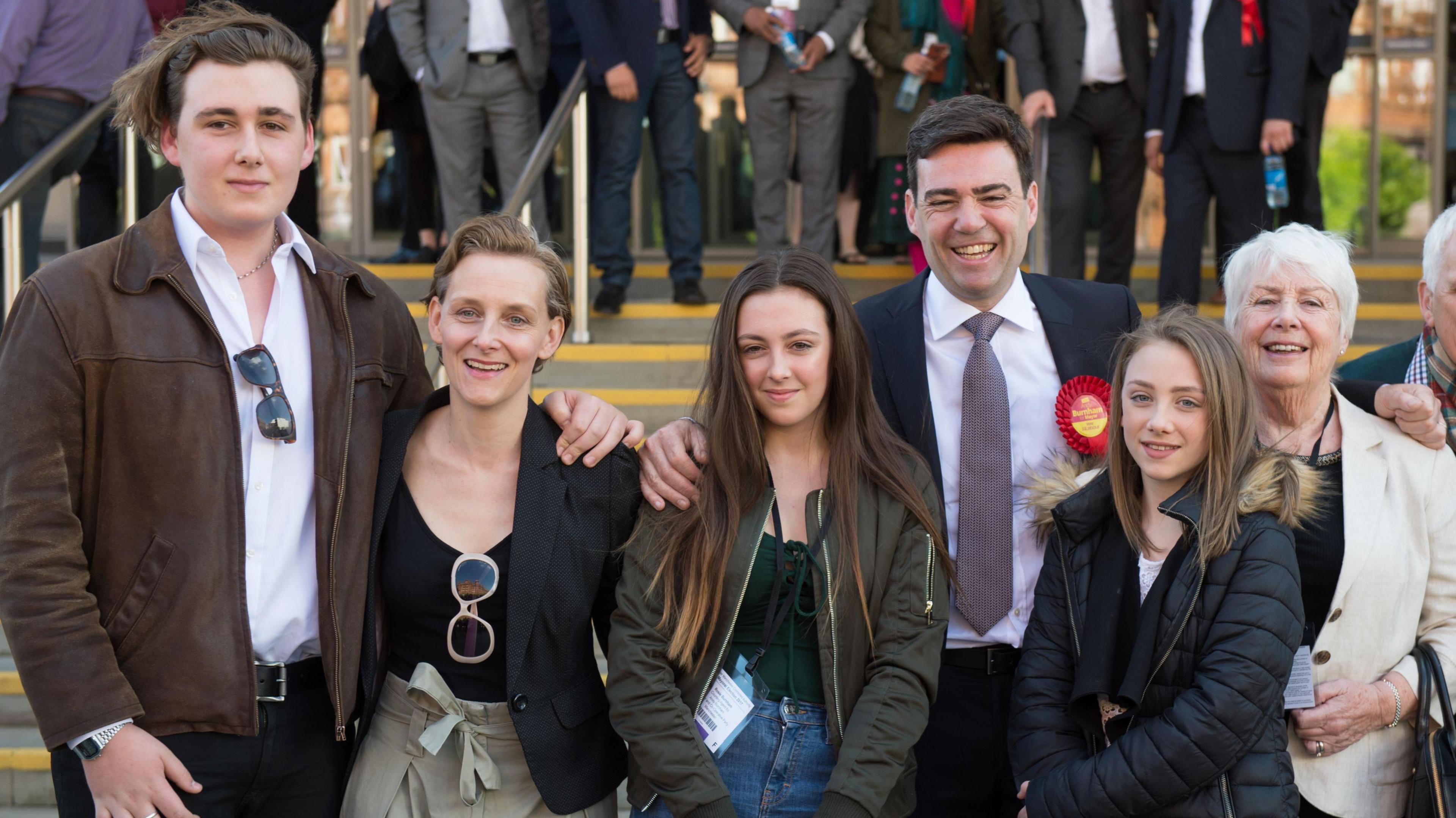 The family look happy and are all dressed casually, standing in a row in front of the convention centre steps. Jimmy is very tall and wearing a brown leather jacket over a white shirt and black trousers, with a pair of sunglasses tucked into the neck of his shirt, and medium length, dark blonde hair held back by a hairband. Van Heel is wearing a tailored black jacket and black scoop-necked tshirt over beige paper-bag style trousers. Her dark blonde hair is worn medium length and slicked back in a quiff. Rosie wears a khaki green bomber jacket over a dark green t-shirt and blue jeans and has waist length, dark brown hair with bleached tips. Annie is wearing a black puffer jacket and a black top and has long, straight, dark blonde hair. Eileen is wearing a cream linen jacket over a black t-shirt and has ear length white hair worn in a pixie cut.