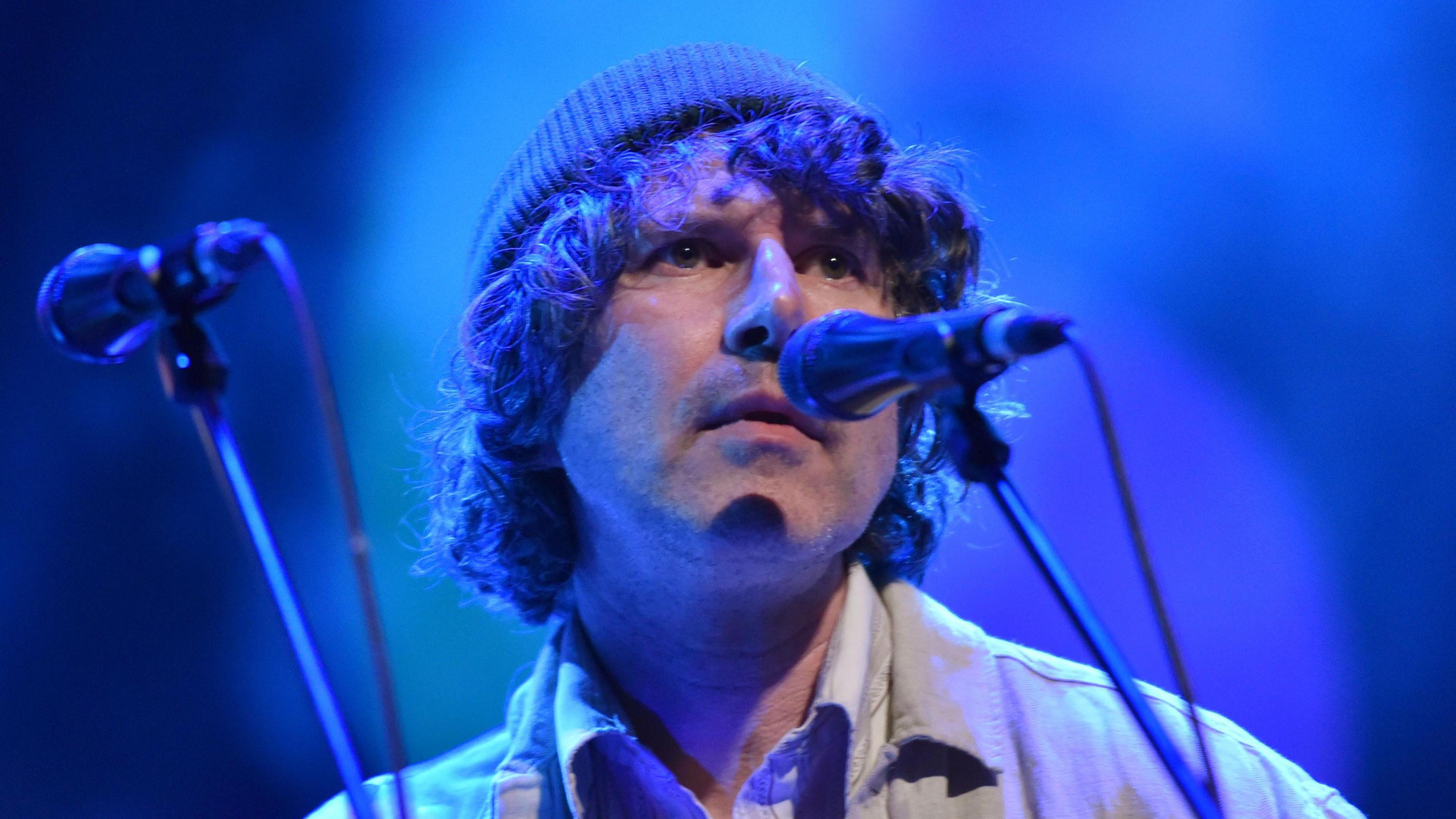 Gruff Rhys performing at 6 Music Festival 2022. He stands on a stage looking out toward a crowd. A microphone on a stand rests in front of his face. He wears a beanie hat and shirt.
