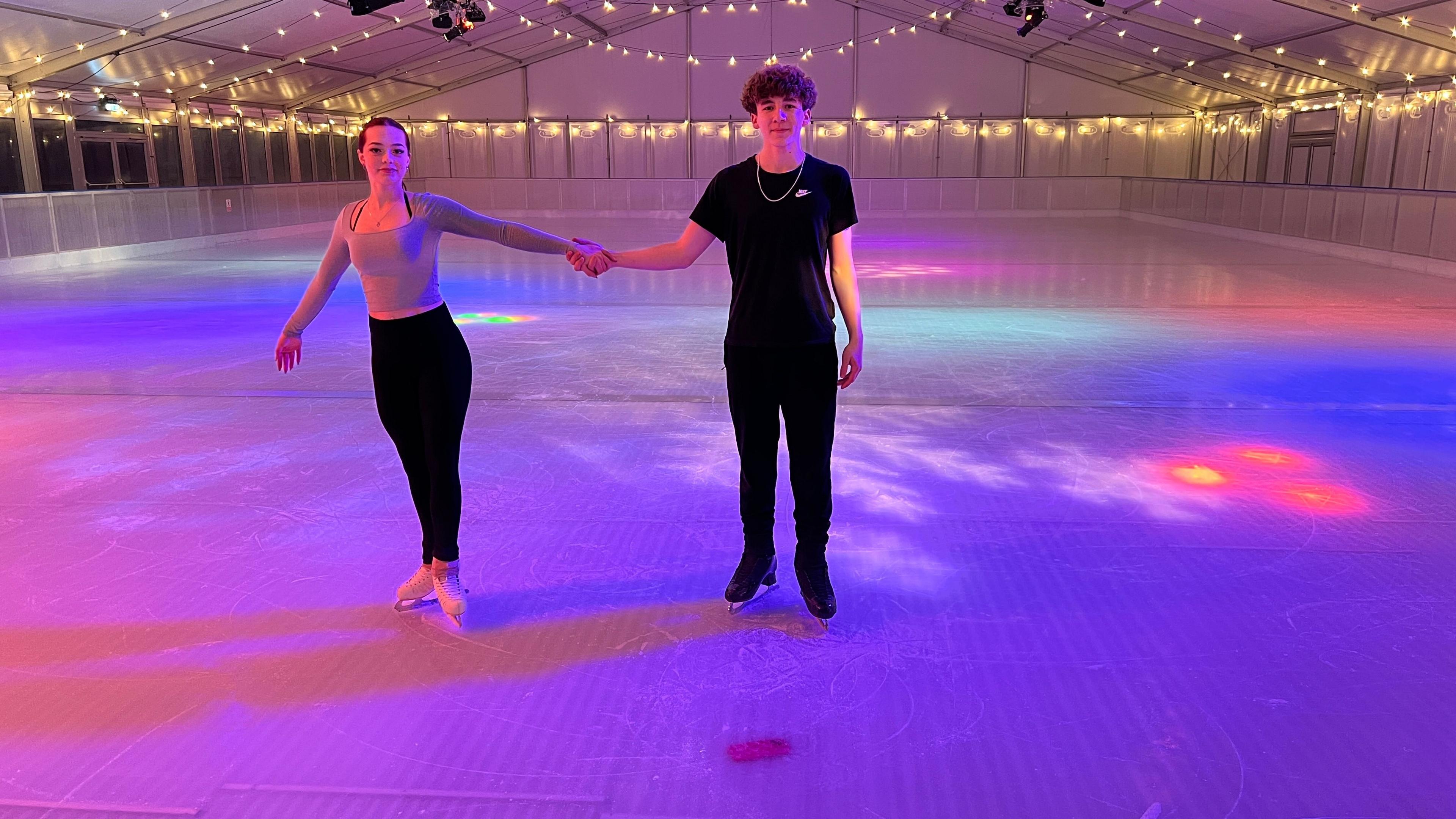 A female skater with brown hair poses alongside a male skater. They are holding hands and looking to the camera. They are on the ice rink and in front of them is the pink smartphone, encased in the ice rink. 