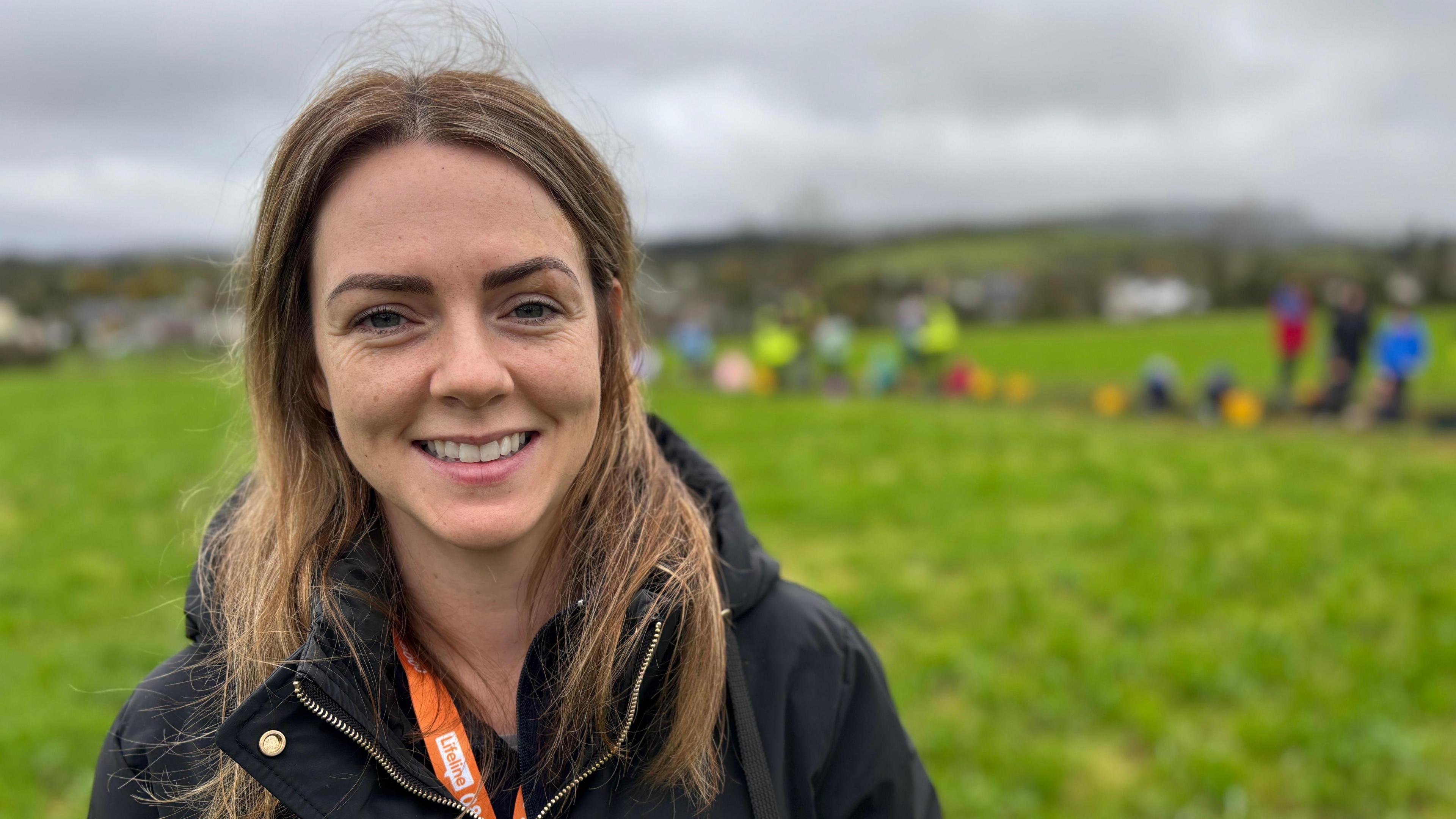 A woman stands in a field. She has long blonde hair and is smiling. She is warmly dressed in a black coat. Behind her, underneath a grey sky is a line of people standing on green grass. They have buckets and are excavating a trench dug into the field. 