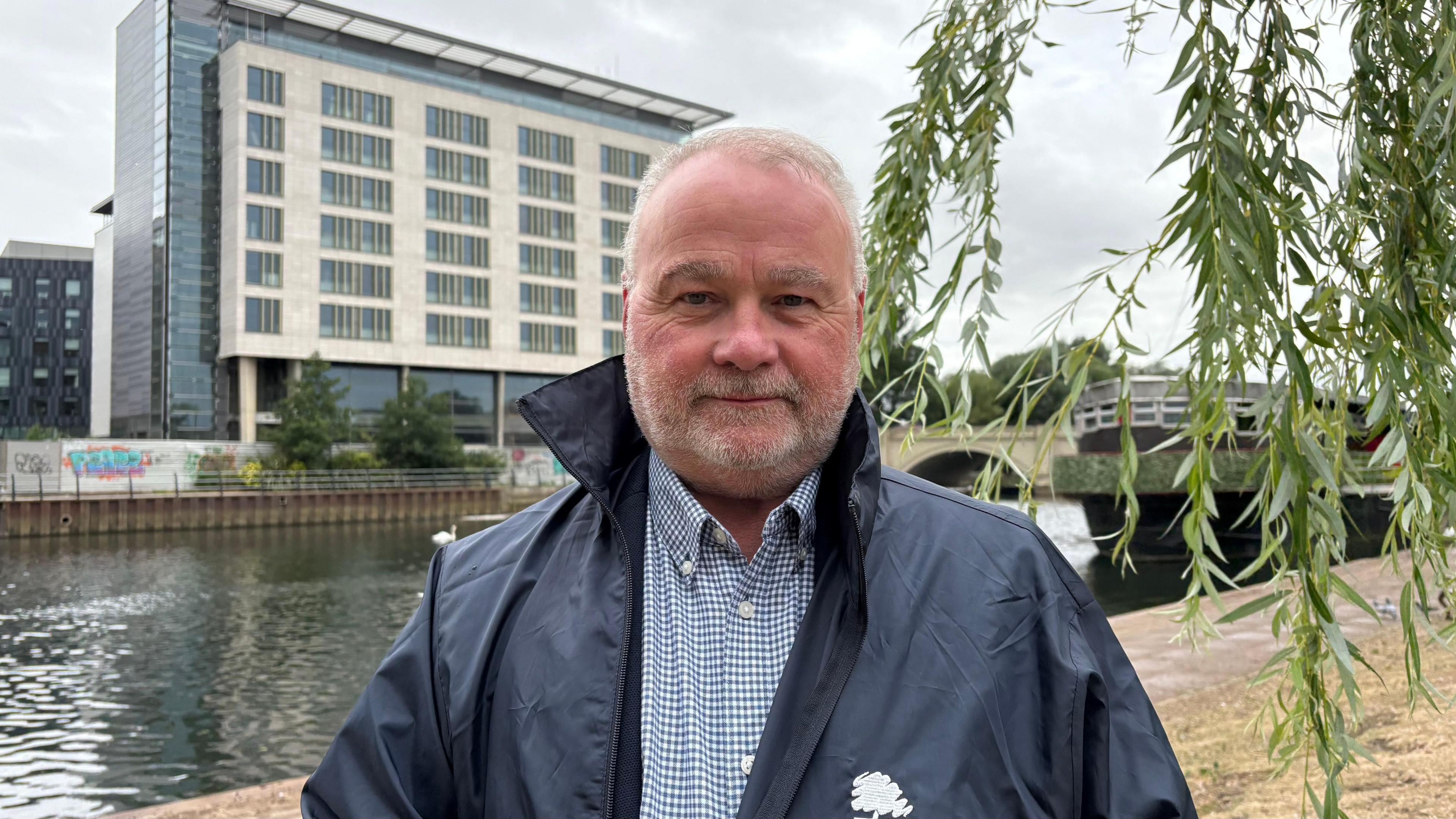 Wayne Fitzgerald looking into camera on the riverbank with the Hilton Hotel in the background. Fitzgerald has short white hair and a white beard. he is wearing a navy rain jacket and checked shirt.
