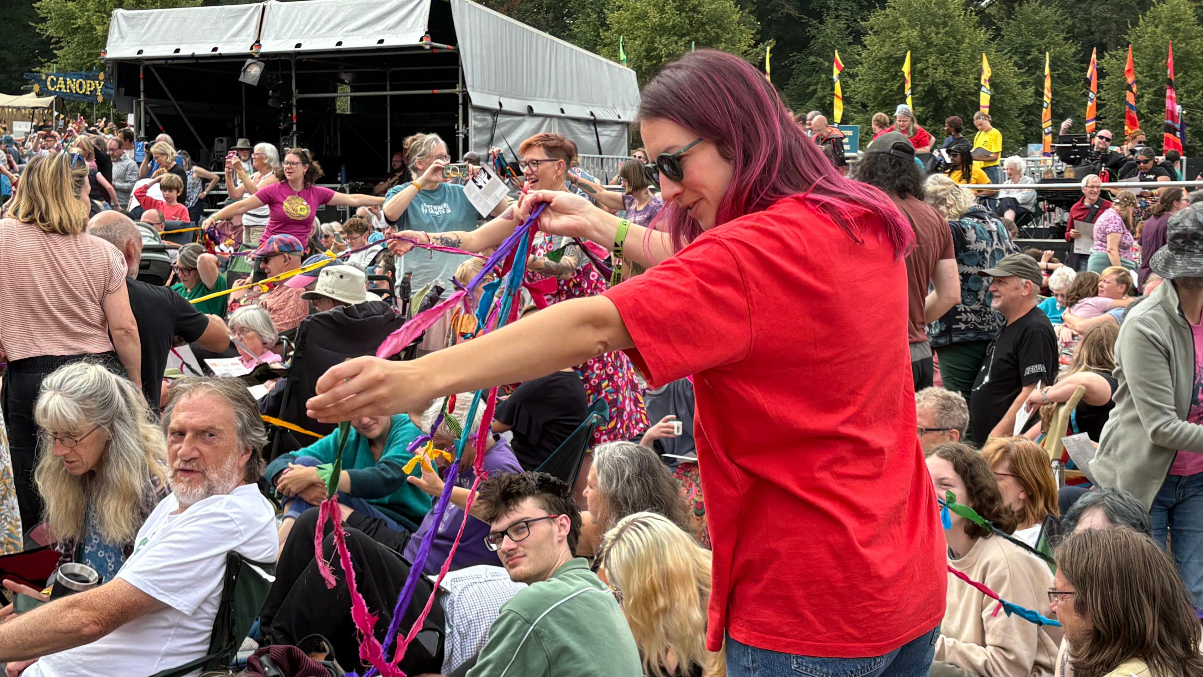 A women with purple hair wearing dark glasses and a red T-shirt is standing in a crown and holding coloured ribbons which are tied together to form chains. There are more chains in the background. There are people sitting and standing in a crowd around her.