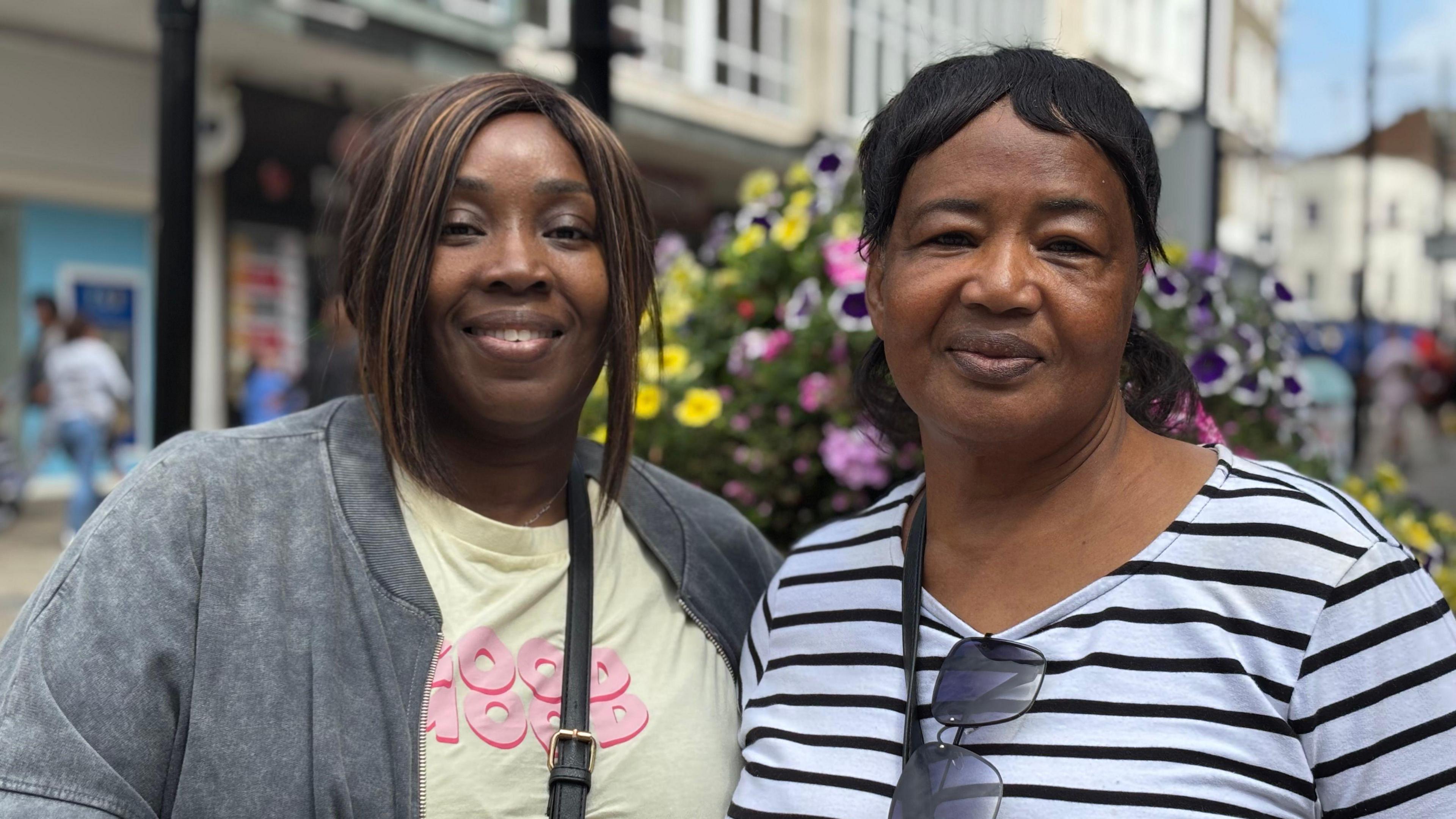 Two women - one in a blue and white striped long sleeved top with a handbag strap over her shoulder and sunglasses hanging off her collar, and the other in a yellow T-shirt and grey jacket with a crossbody bag strap visible, are standing on a high street and smiling at the camera. There are shops, floral displays and people blurred in the background.
