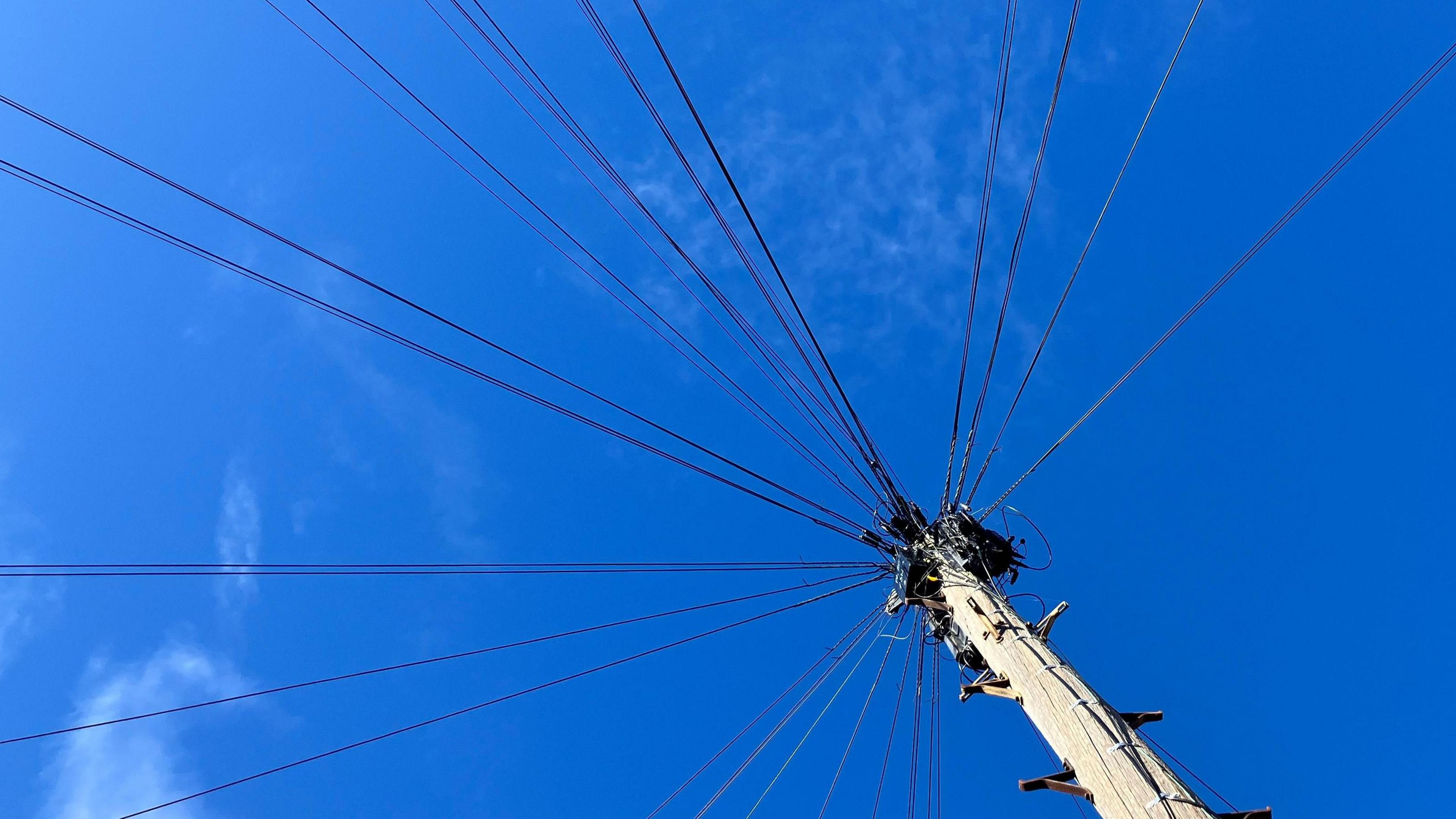Taken from the bottom of a telegraph pole looking up towards blue skies with numerous wires heading out in different directions at the top and climbing pegs sticking out of the wooden pole itself.
