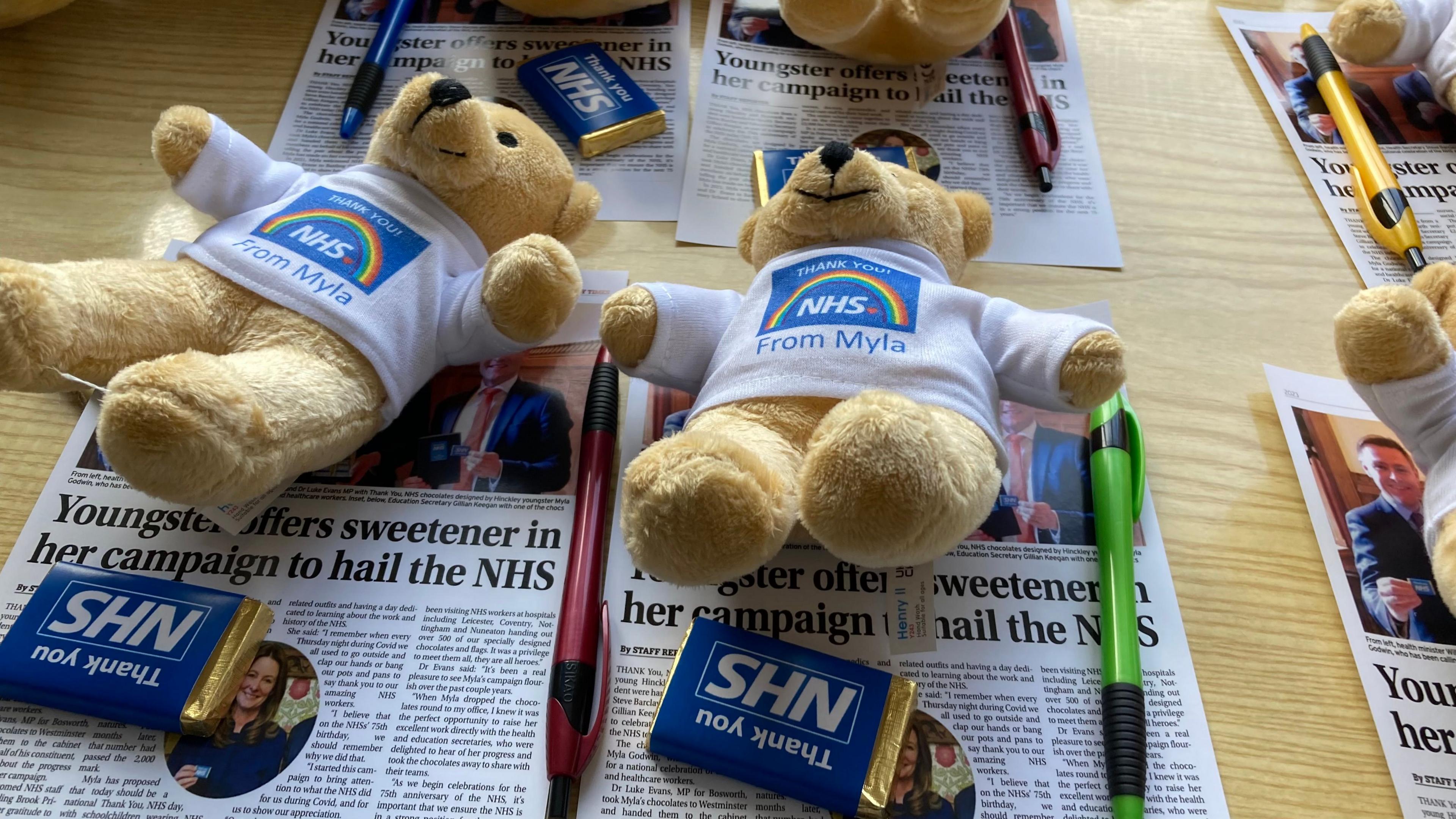 Yellow teddy bears with white t-shirts on with a NHS logo that reads 'thank you from Myla'. They are laid on top of leaflets. There are also colourful pens on the table and cubes of chocolate labelled 'thank you NHS'.