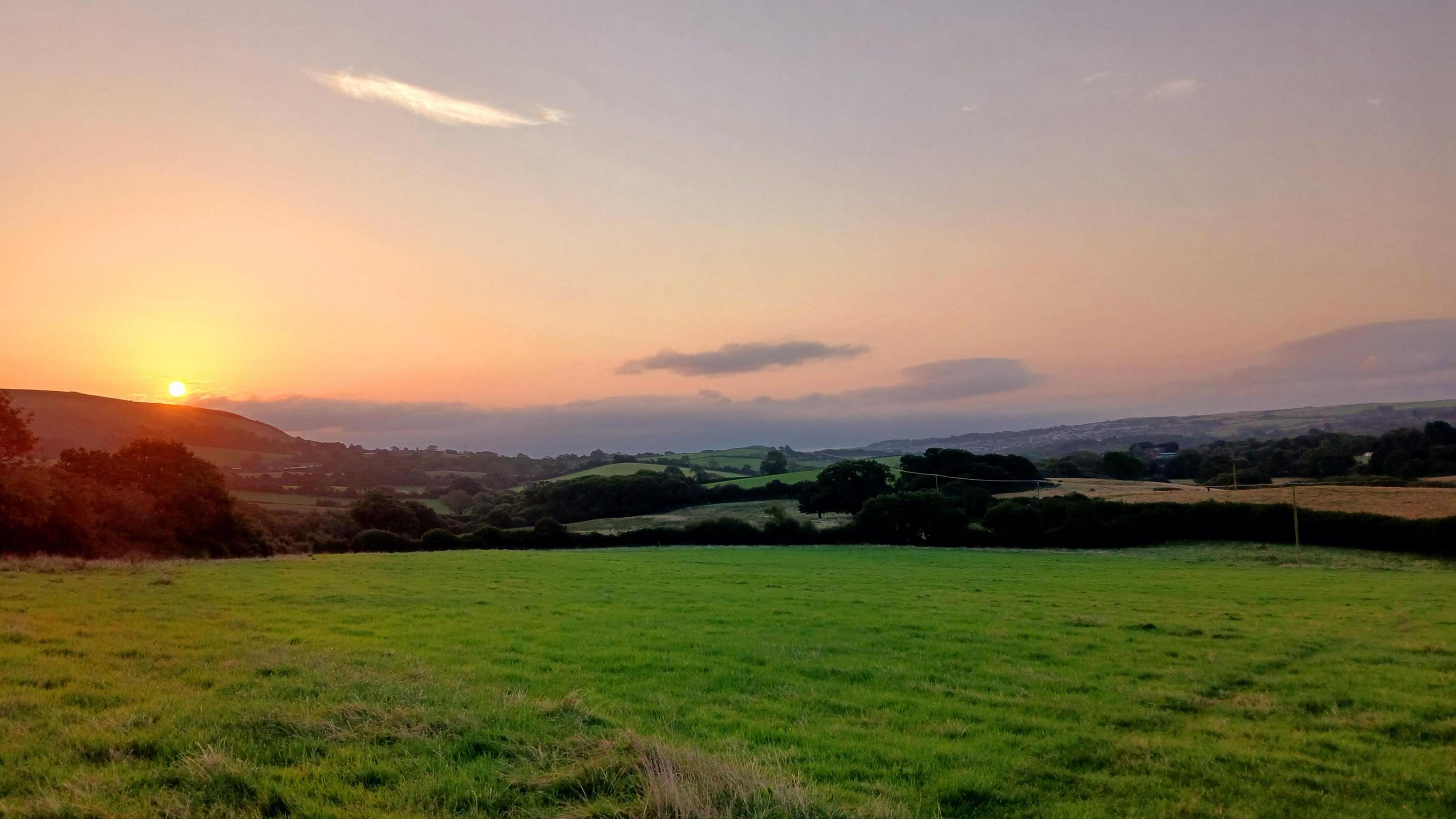 Rolling green fields can be seen up to the horizon under a low-lying sun and with just a few clouds in the orange-blue sky. 