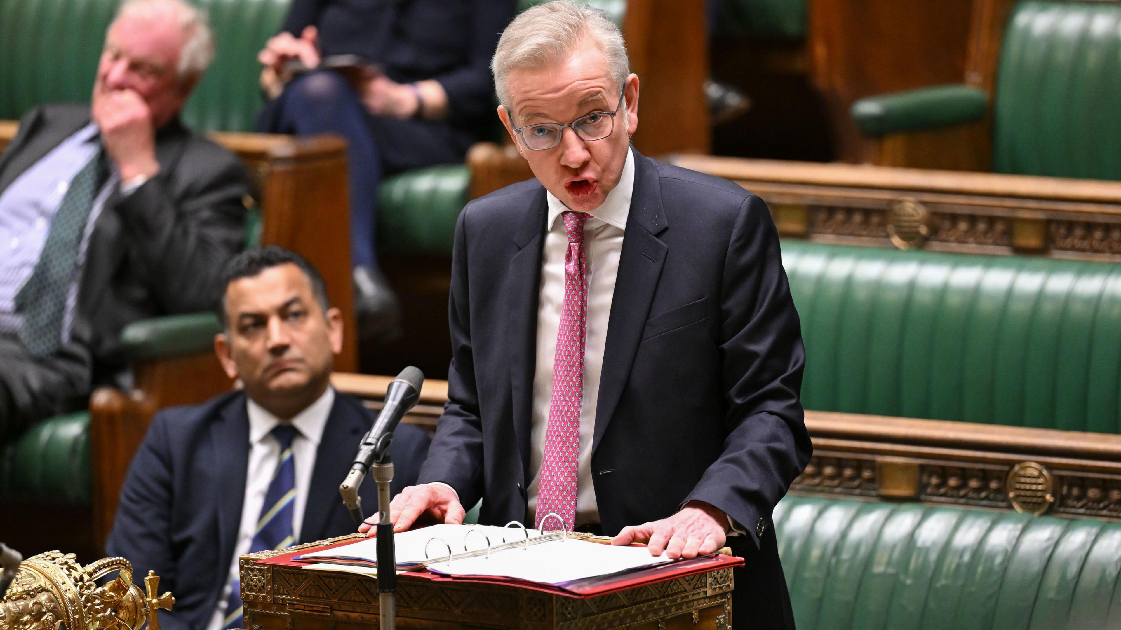 Michael Gove stands at the dispatch box in the house of Commons, talking to the House. He is wearing a navy suit, white shirt and pink tie.