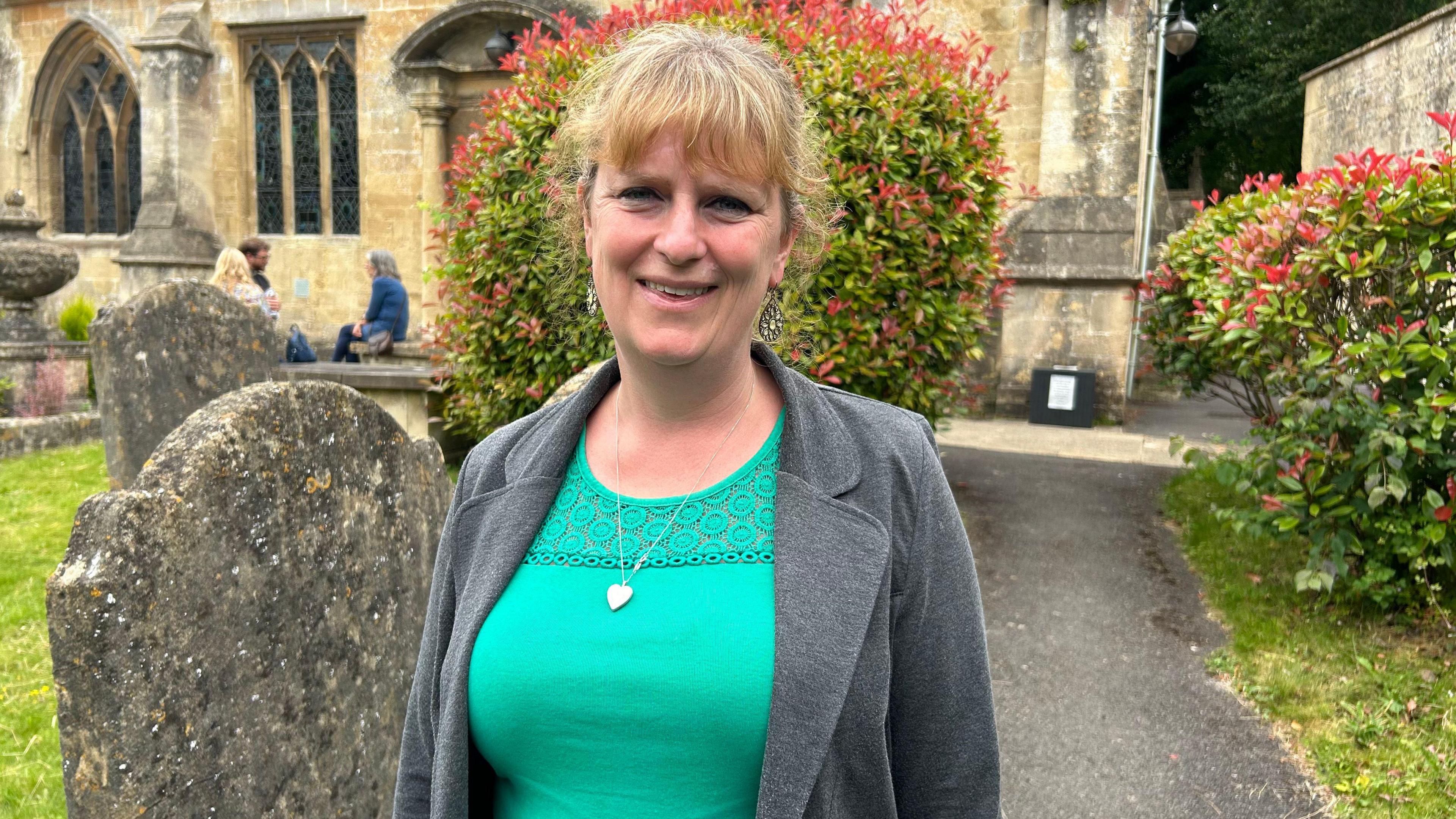 Emily Aspin stood outside of a church wearing a grey blazer, green top and silver heart necklace