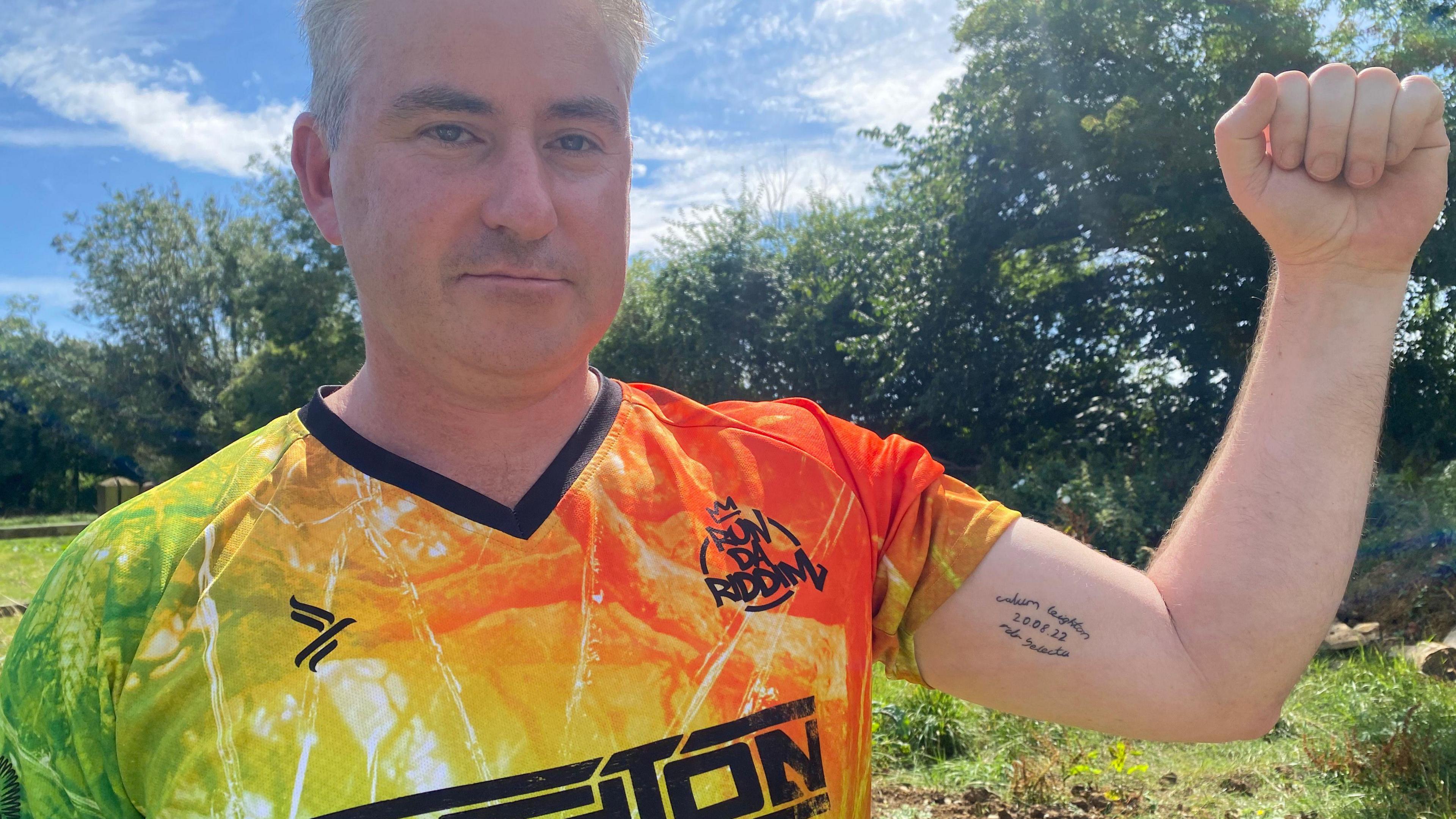 Andrew Leighton wears a brightly coloured top featuring the record label's logo. One sleeve is rolled up to show off a tattoo on the inside of his left arm.