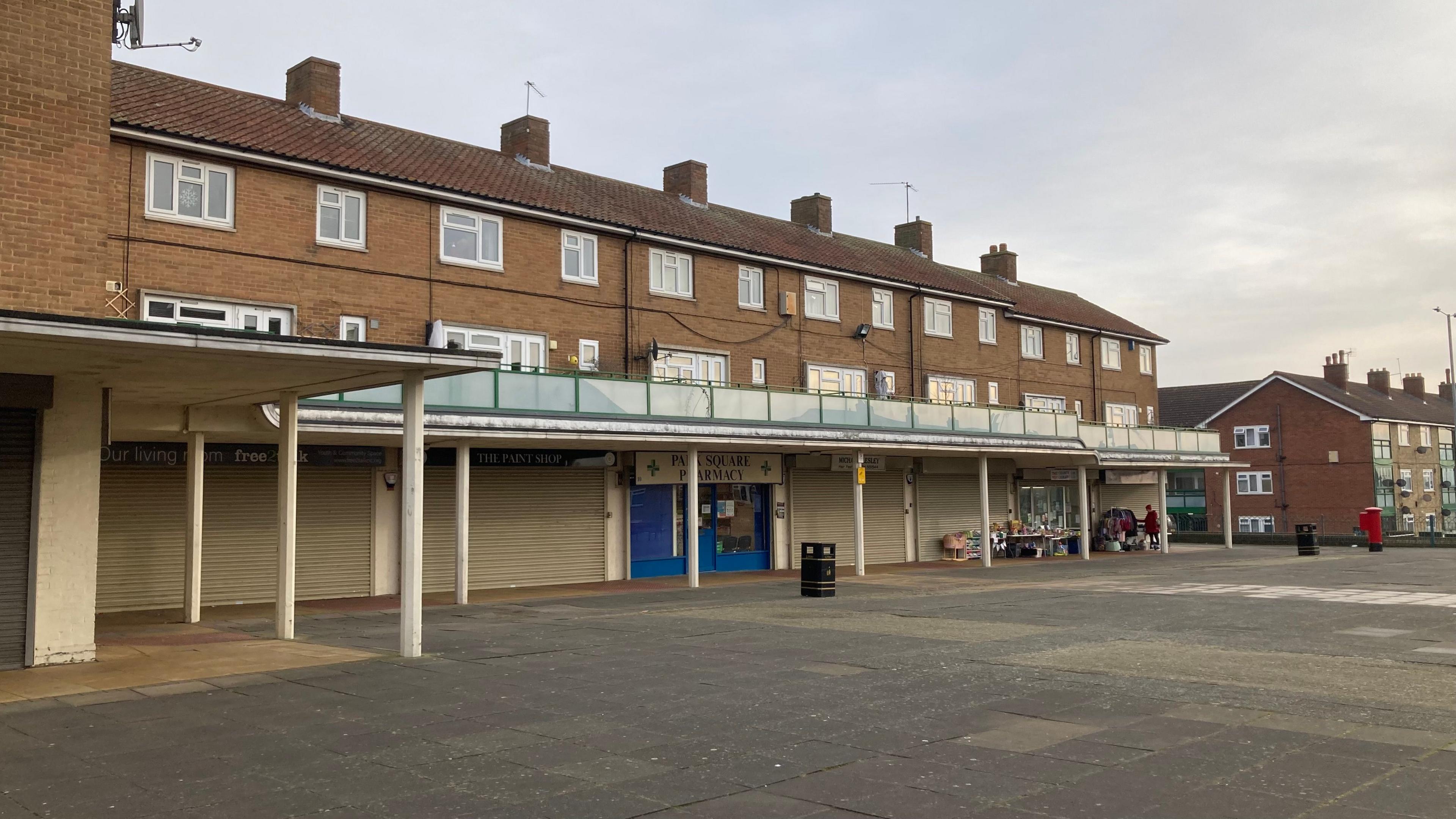 Large slabbed area, with a parade of shops. Most are empty and covered by shutters. There is a blue and white pharmacy unit in the centre, and a toy library with toys on tables at one end of the parade.  There are pillars outside the shops which support the walkway serving the flats above.