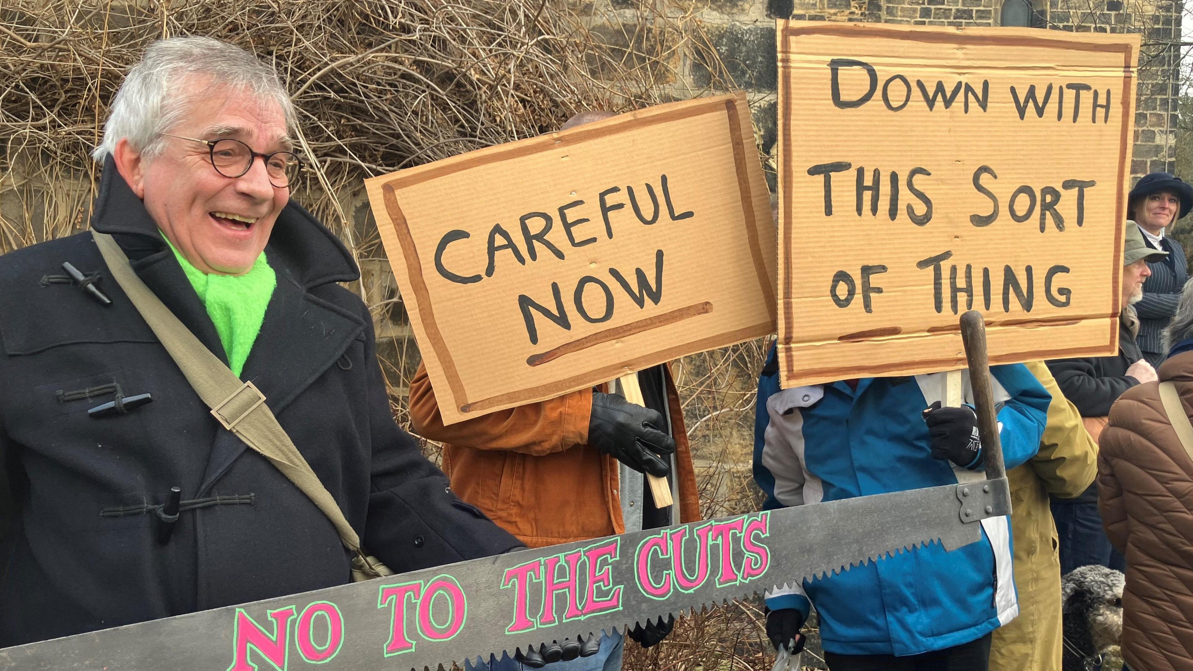 A grey-haired man wearing black-rimmed spectacles, a black duffel coat and a green scarf stands next to two people holding placards in front of their faces. The placards read, "careful now" and "down with this sort of thing".