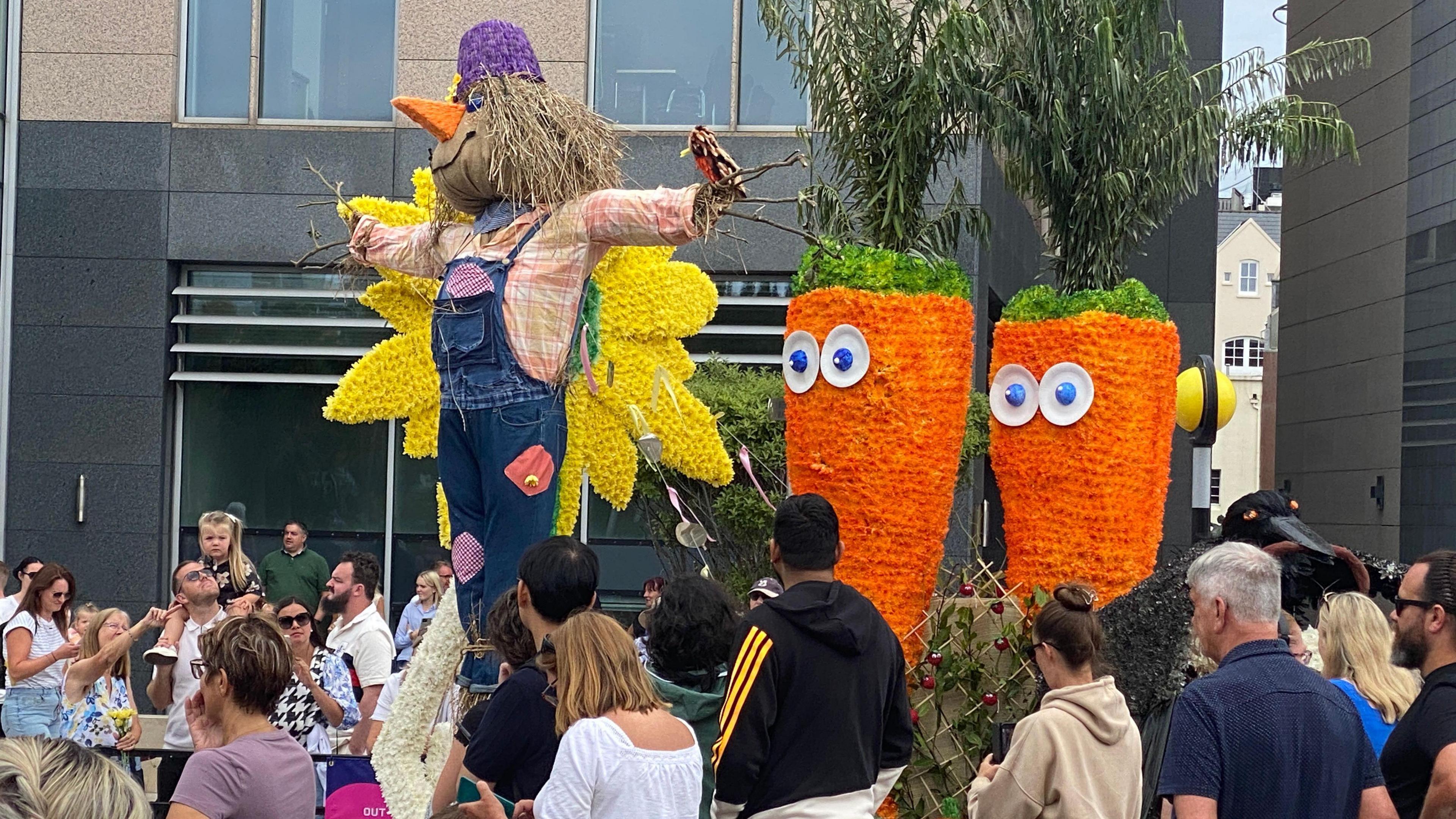 A float which features a giant scarecrow wearing patched dungarees. It has a carrot nose and a bird crafted out of sticks sitting on one of its outstretched arms. There are two giant carrots which have palm trees as their green leaves and a pair of eyes on the front of them. Behind the scarecrow is a giant sunflower. Spectators are looking up at the float.