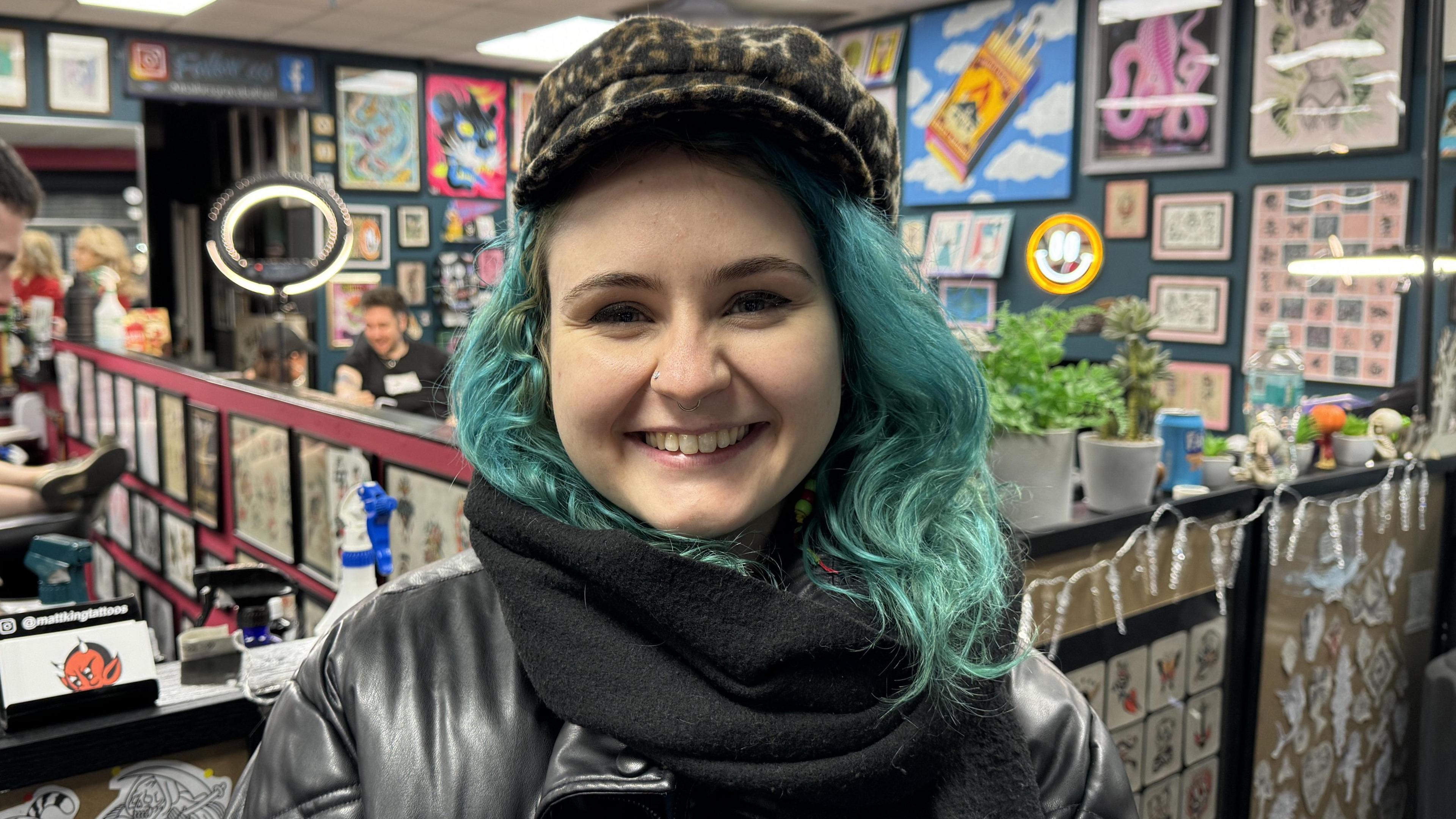 Hannah wearing a leopard print hat, black scarf and black leather jacket, she has blue hair 