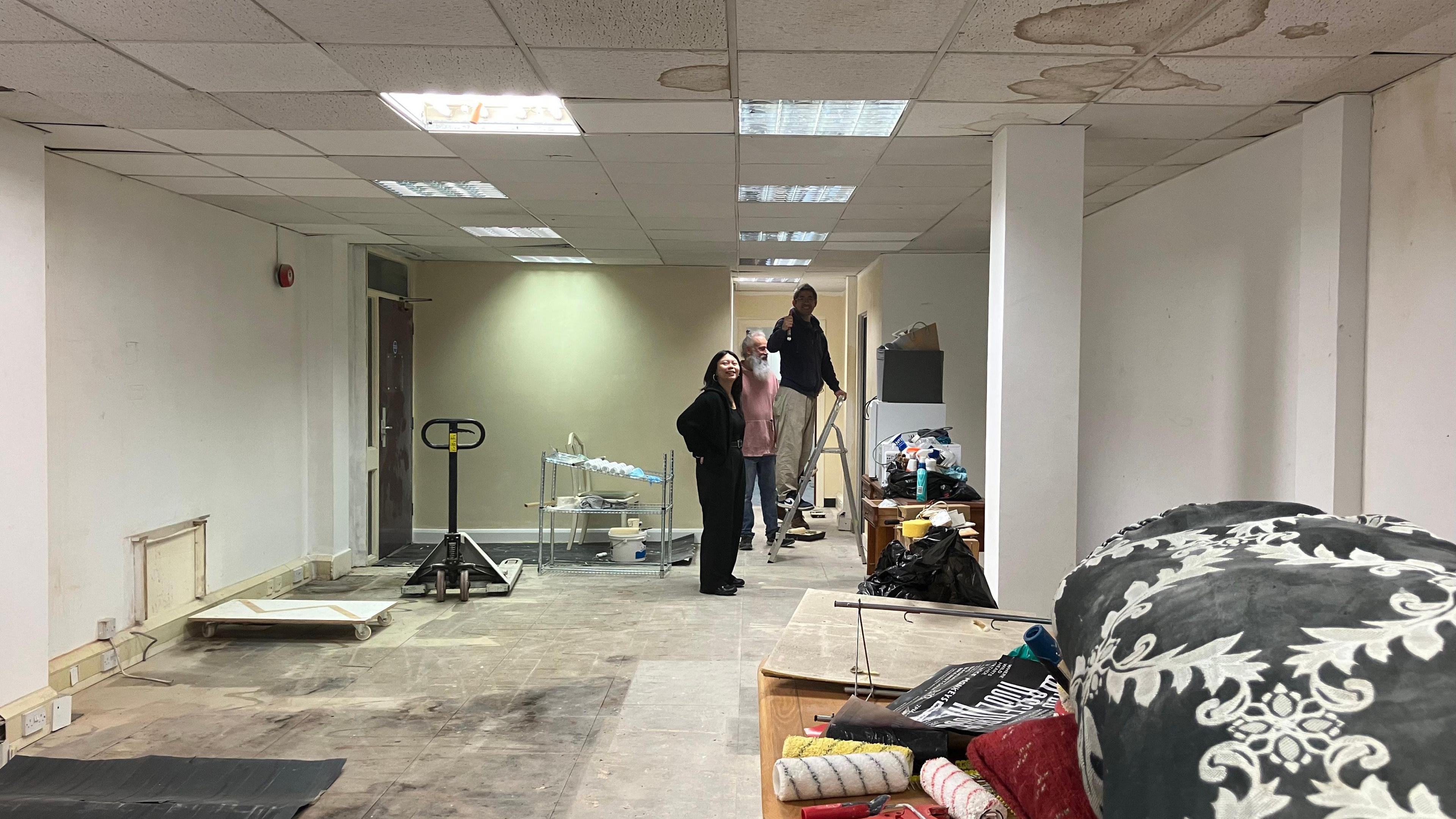 The inside of a mainly empty shopping unit, with no floor tiles and stains on the ceiling tiles. There are several people stood at the end, one is on a ladder. there are paint rollers in the foreground