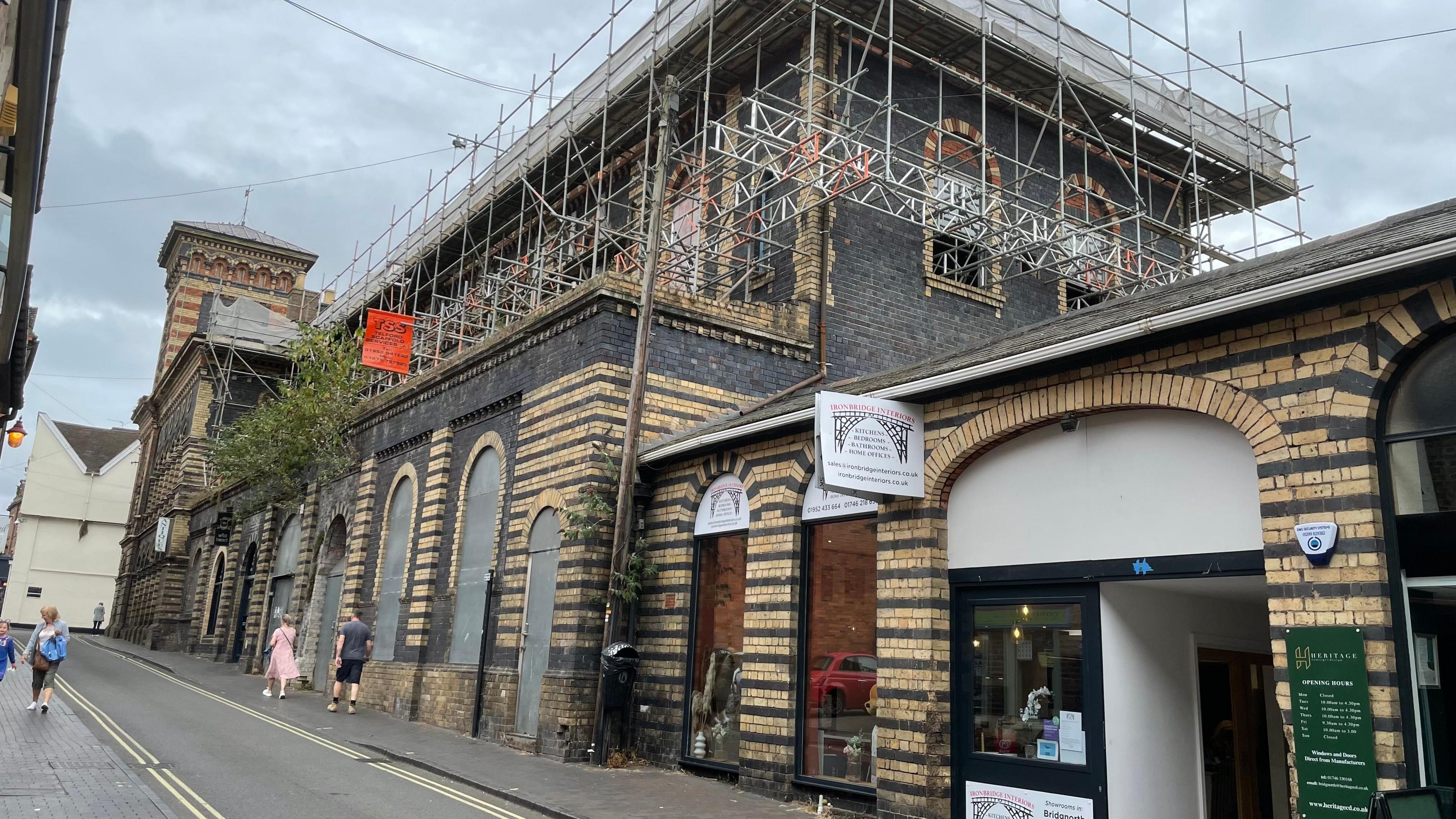 Scaffolding erected around the New Market Buildings, which has vegetation growing from outside the first floor walls. Windows and doors can also bee seen boarded up