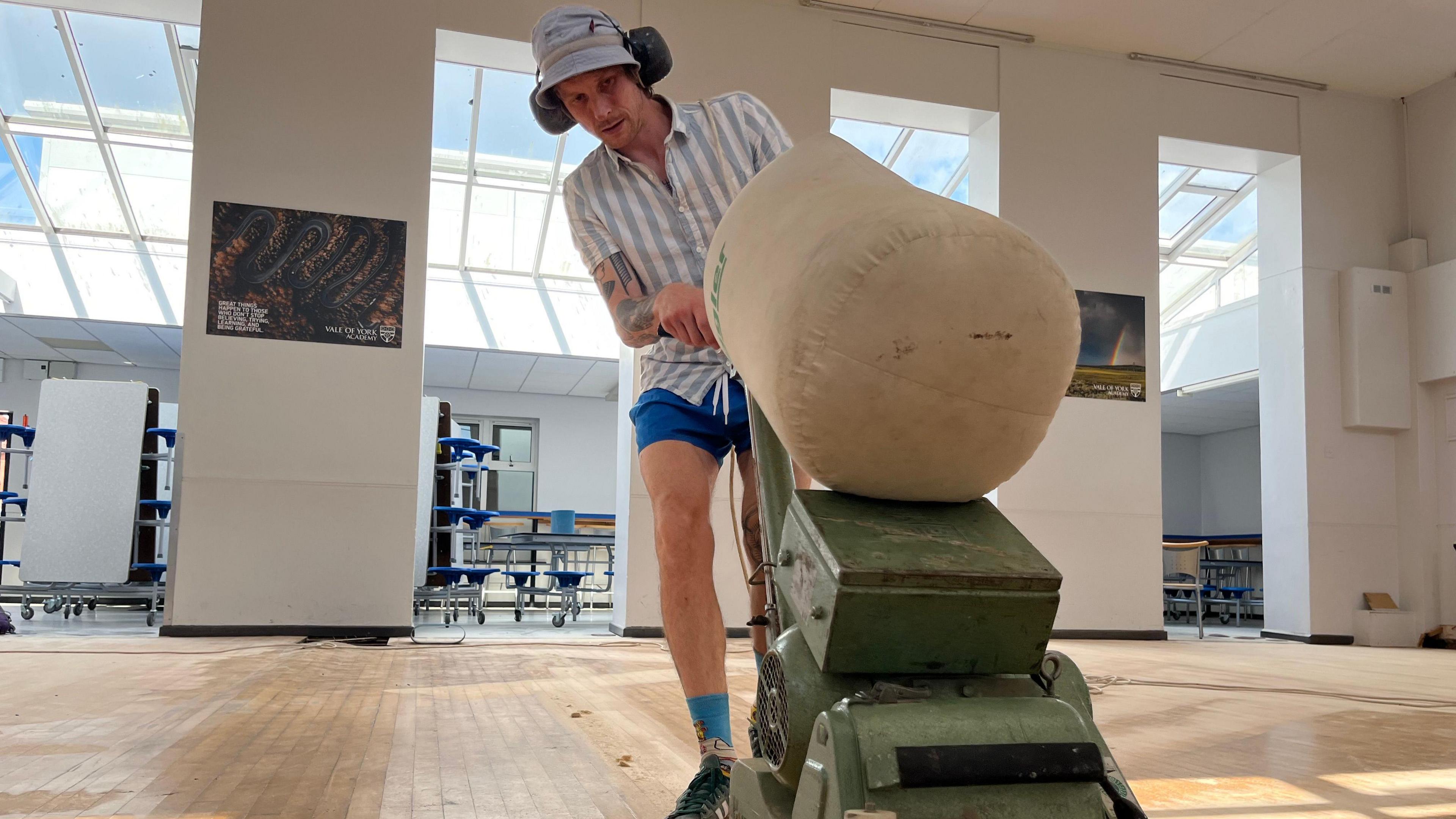 Danny Stenhouse wears ear defenders and a blue shirt and shorts as he pushes a large floor sander across a wooden floor