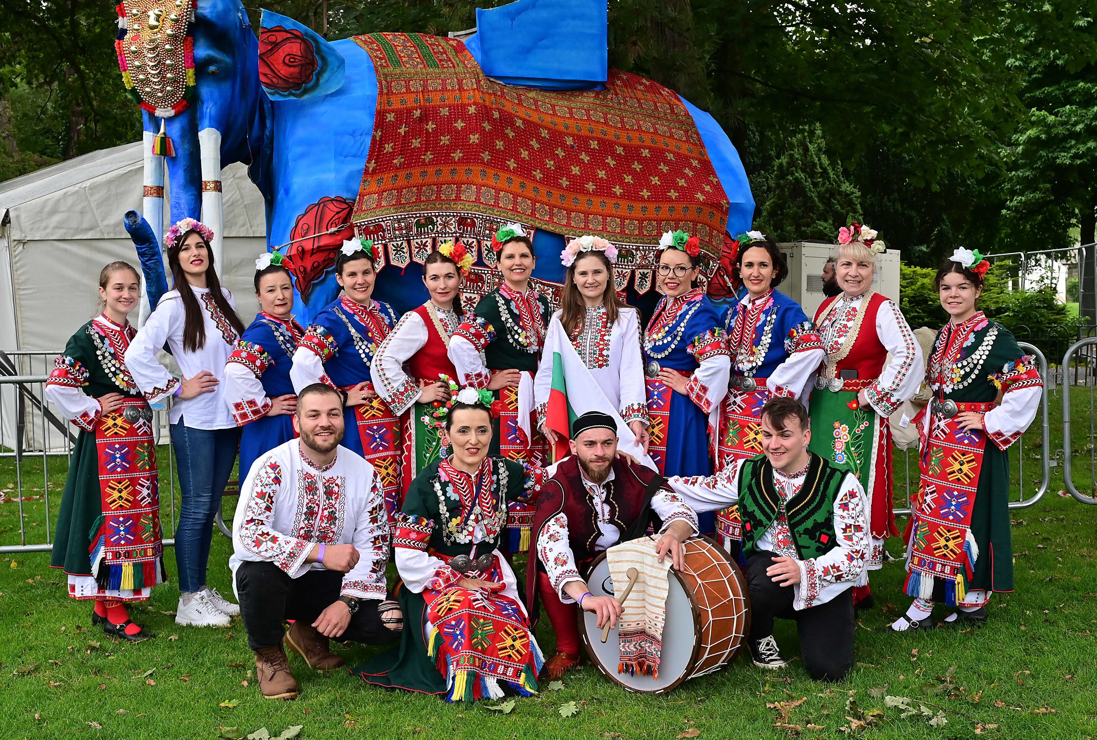 A large blue elephant puppet, life sized, with ornate red blanket on it, a large group of people pictured smiling in front of it, all dressed in bulgarian folk dress featuring red, white and green patterned clothing