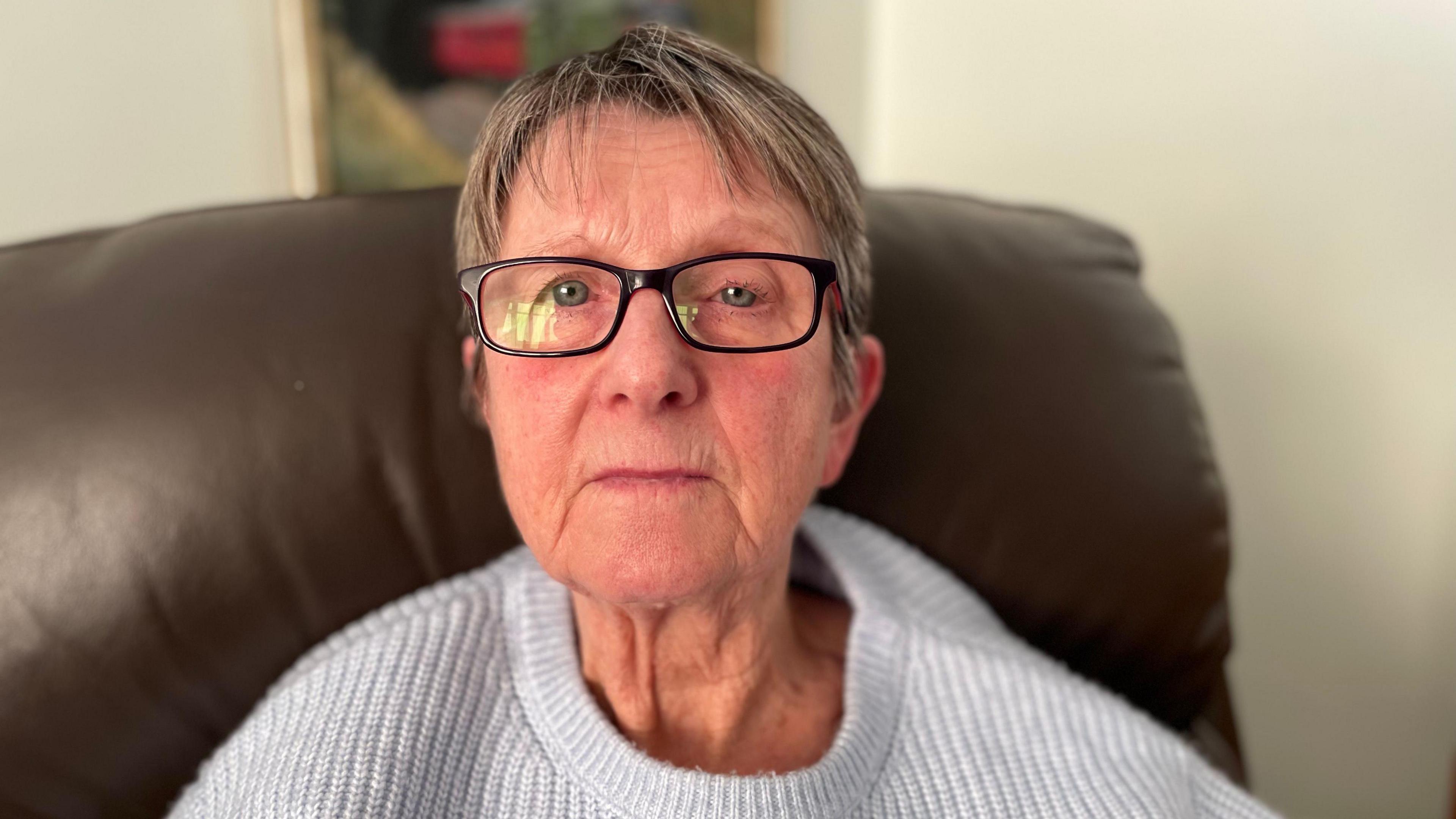 Woman in her 70s with glasses and a light-coloured jumper sitting on a brown leather sofa