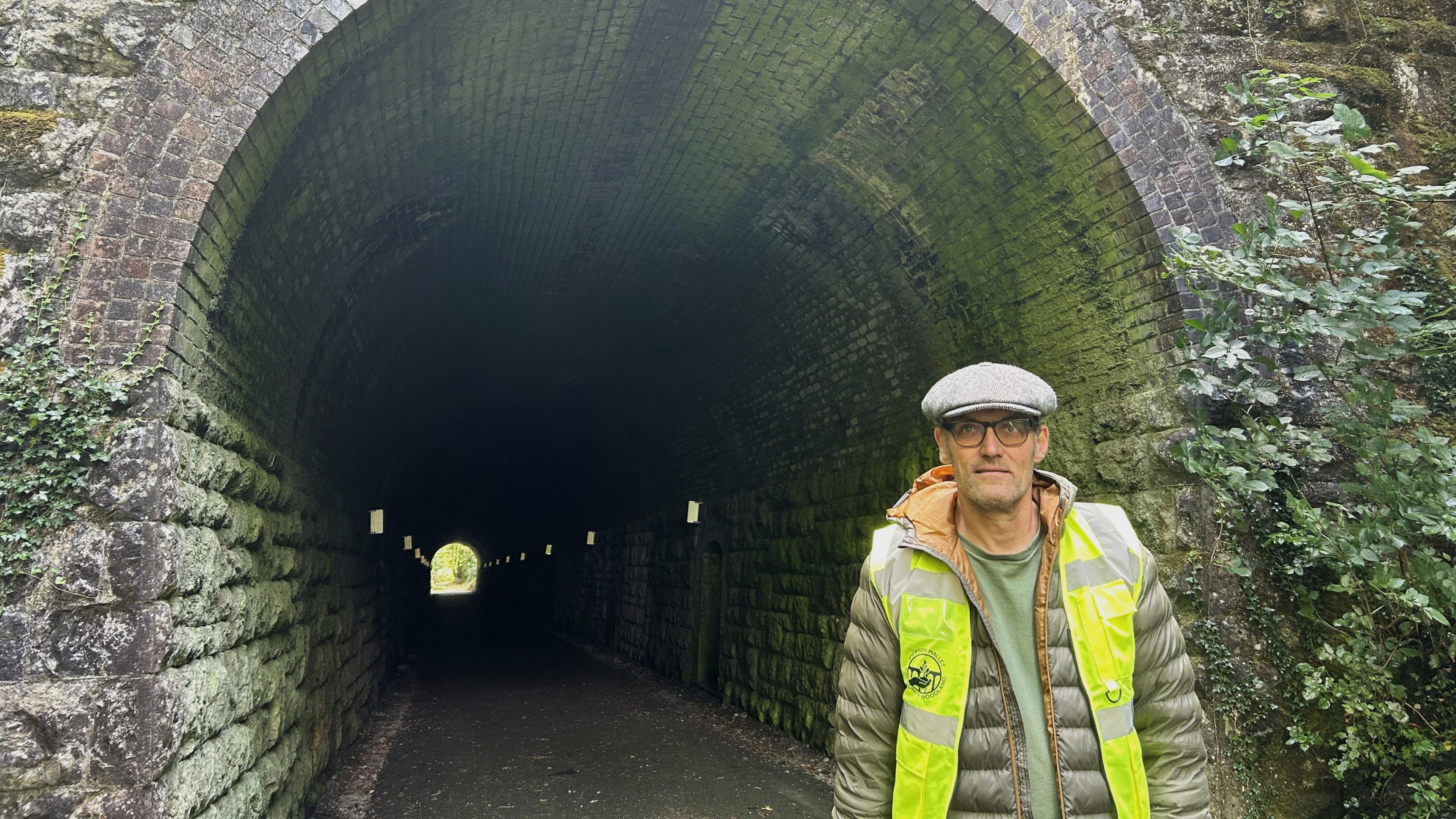 Gavin Mayall wearing a pale green padded jacket underneath a yellow high-vis jacket, a grey cap and tinted glasses. He is standing at one end of the Windsor Hill Tunnel, a large stone arch with ivy growing up the sides.