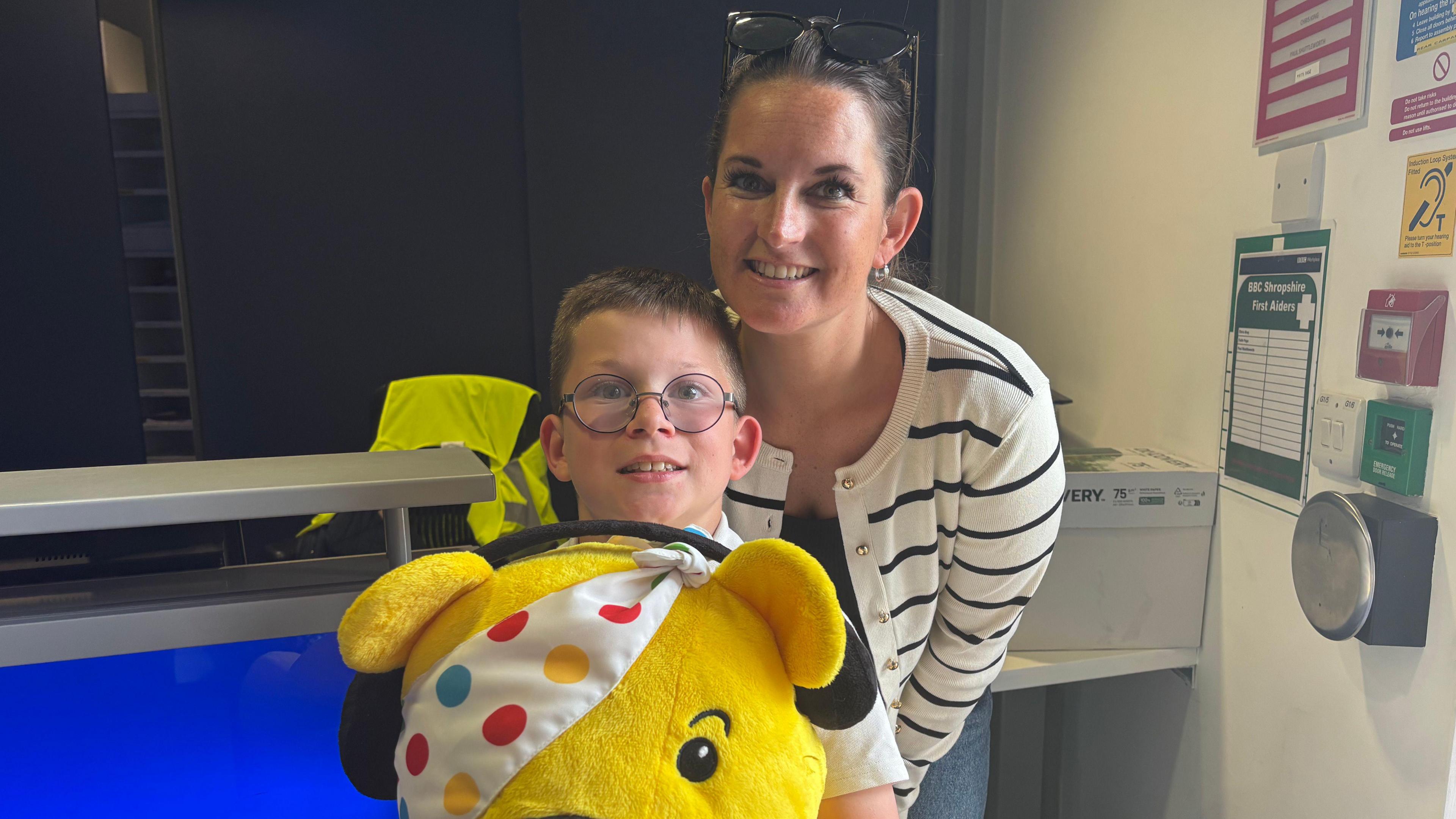 Oscar is wearing glasses and holding a yellow Pudsey bear plush toy with a spotty eye patch. His mum, Steph, has dark brown hair tied back, with sunglasses on his head.