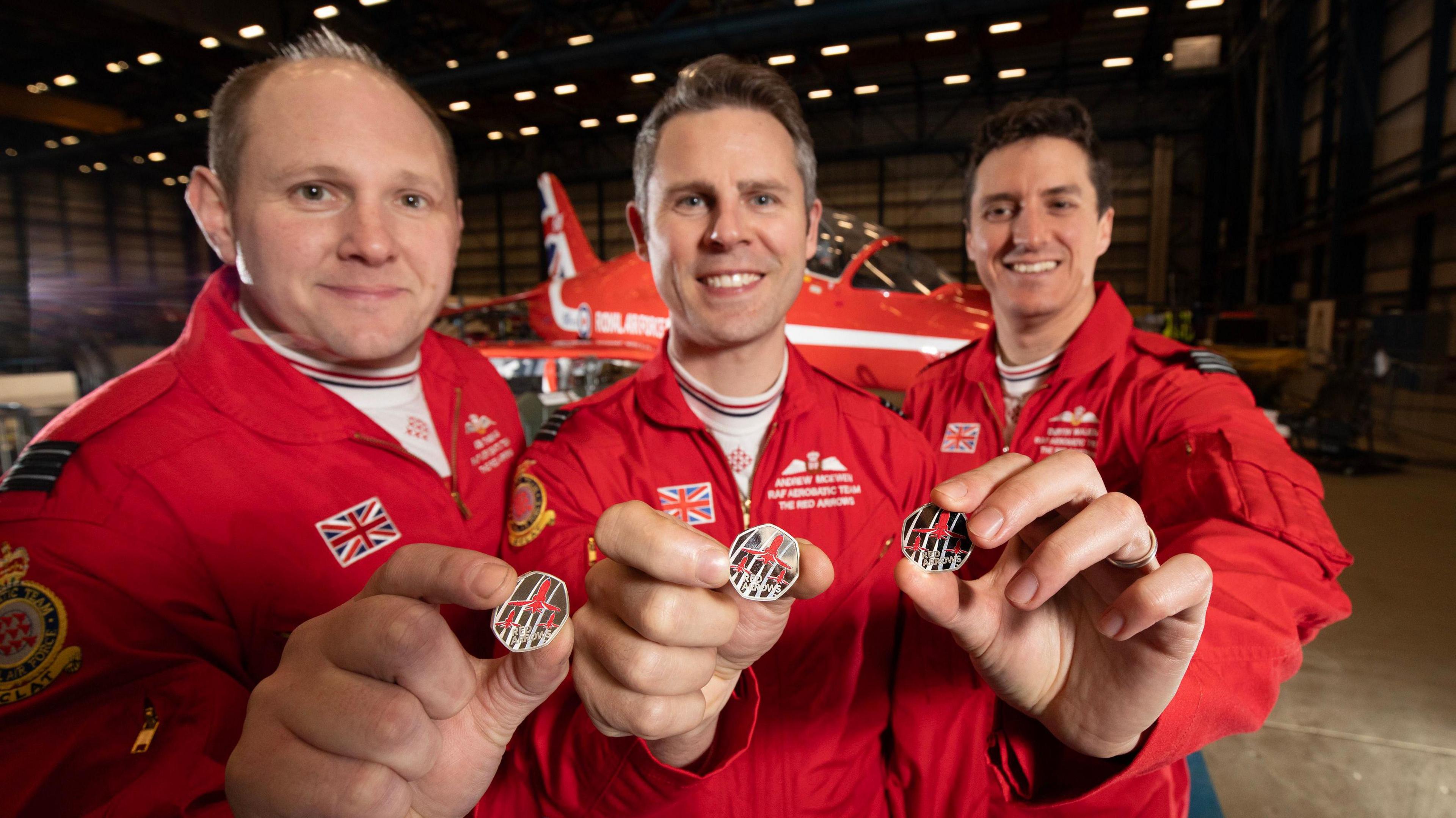 Three clean-shaven men with short hair wearing red RAF overalls. They are smiling and each holding up a Red Arrows 50p coin. There is a Red Arrows plane in the background.