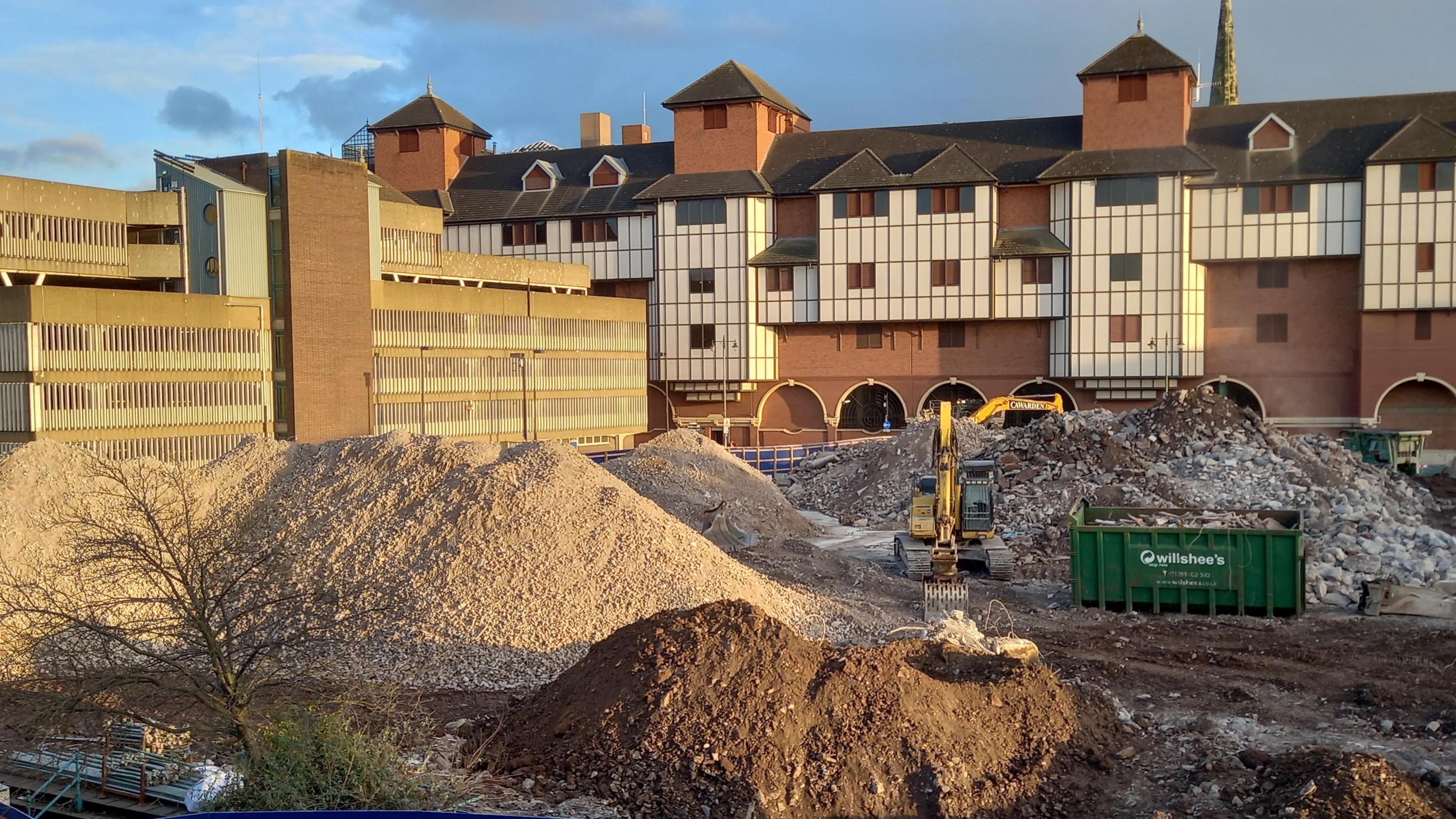 A building site with a yellow digger among large mounds of rubble and earth, after a building has been knocked down. Multi-storey buildings can be seen to the side and behind the building site.