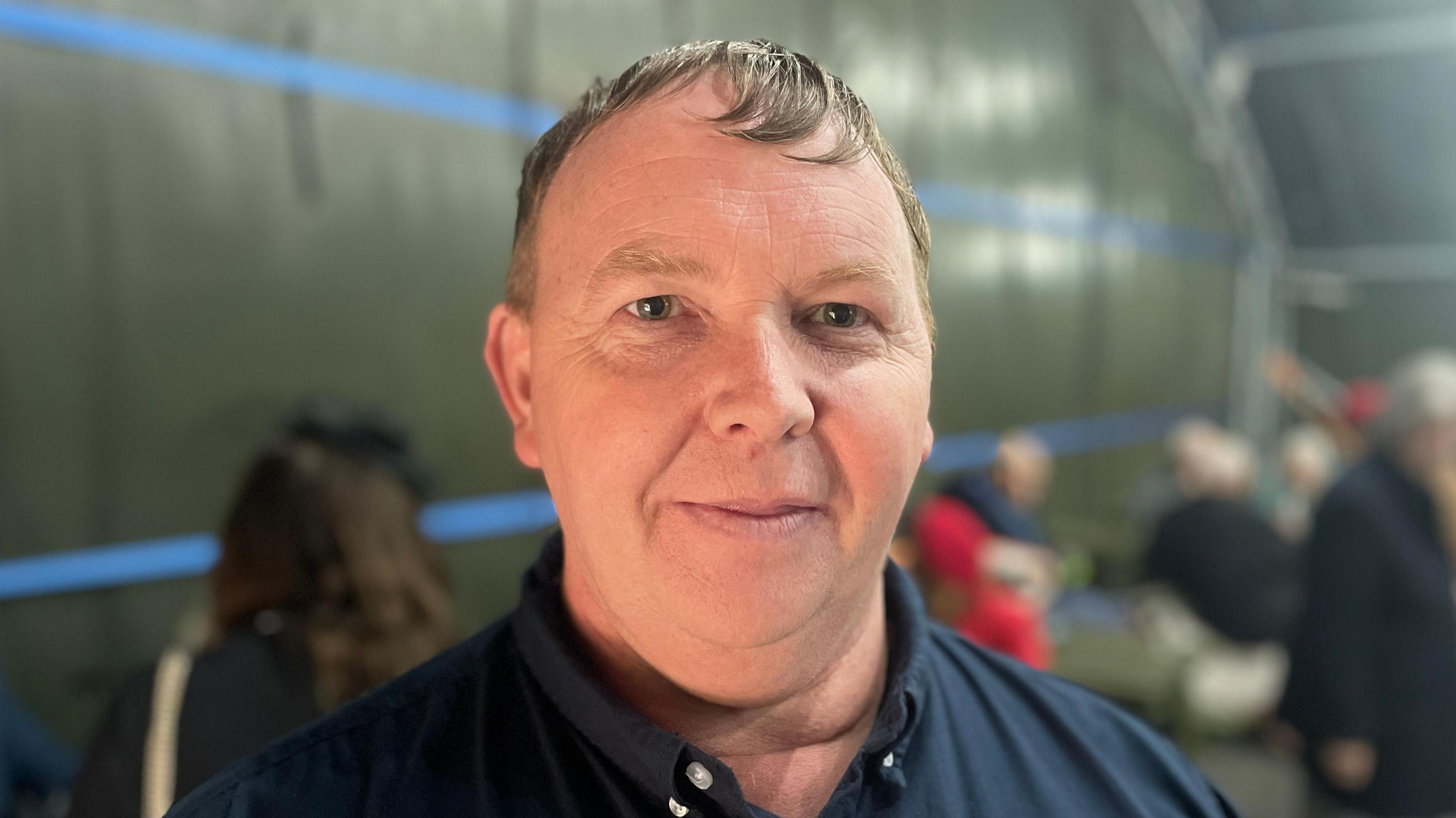 Andy Smith wears a navy blue polo shirt and looks into the camera. He is standing in a former air shelter which is being used as the venue for Dorothy Smith's birthday party