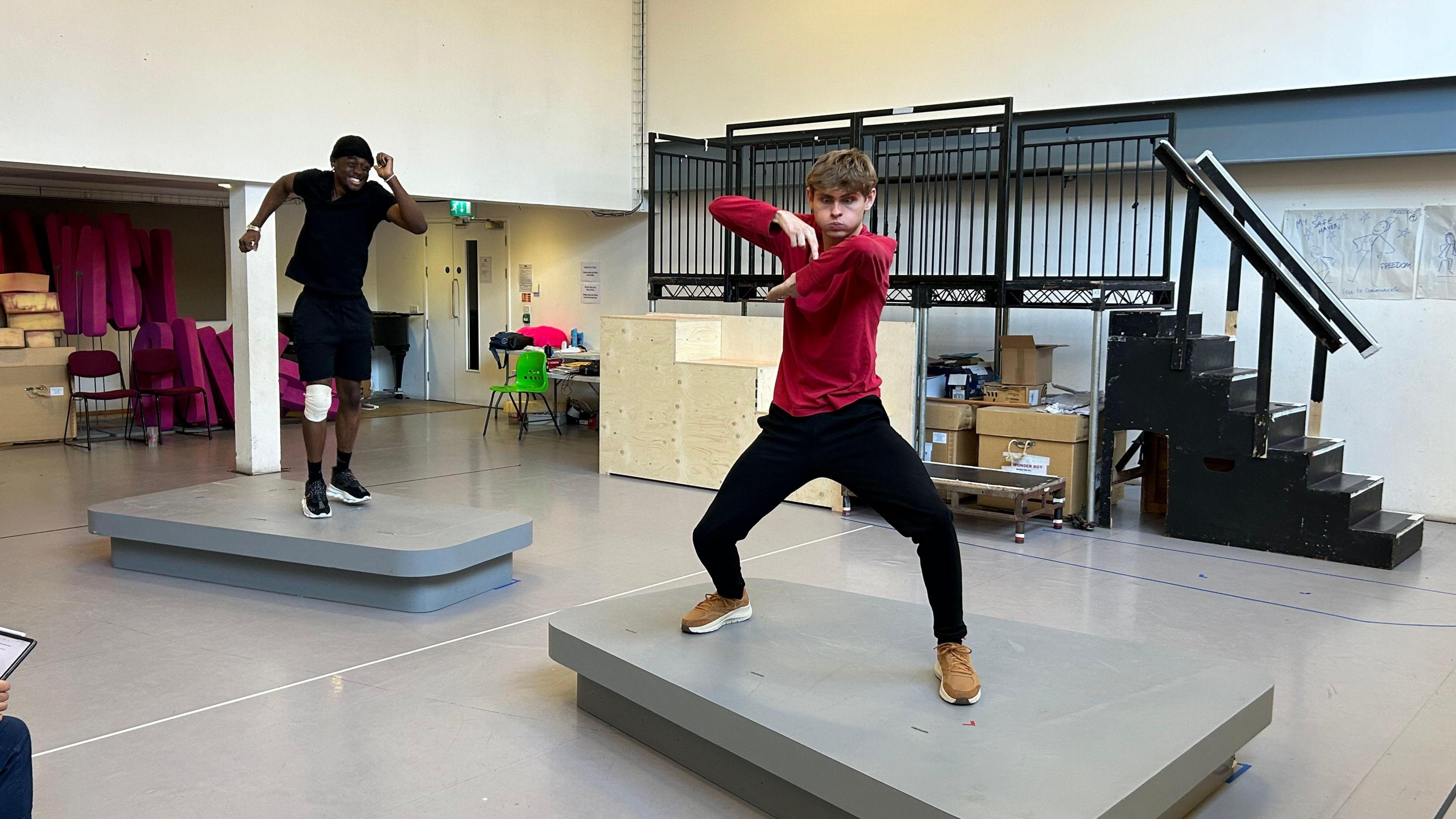 Ciaran O'Breen in a red long-sleeved top and black trousers making gestures with his hands while standing on a platform. Another actor, dressed all in black, is standing on a platform to the left, looking like he is running