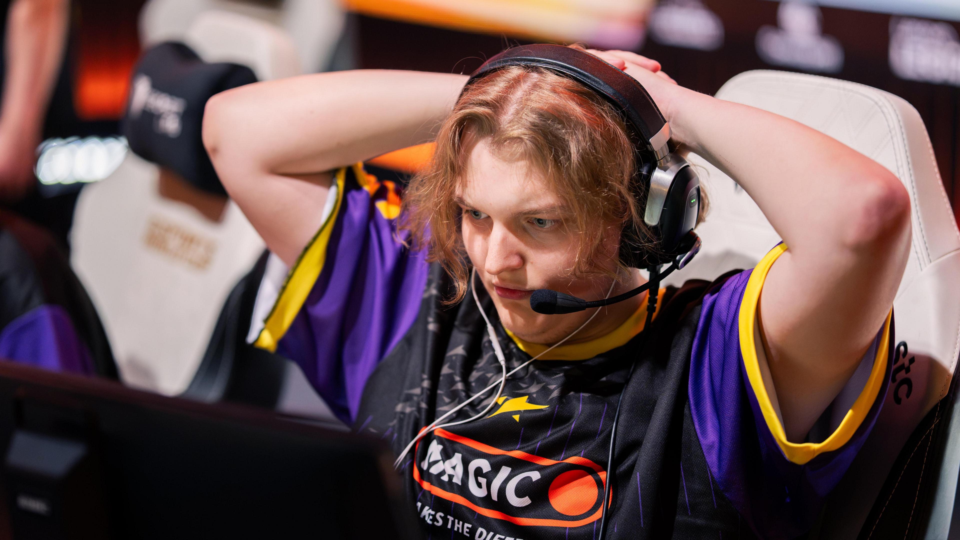 A competitor at the Esports World Cup watches a screen intently with their hands clasped behind their head, creating an impression of anticipation or nervousness. They wear a large gaming headset with a microphone and a black and purple team jersey.