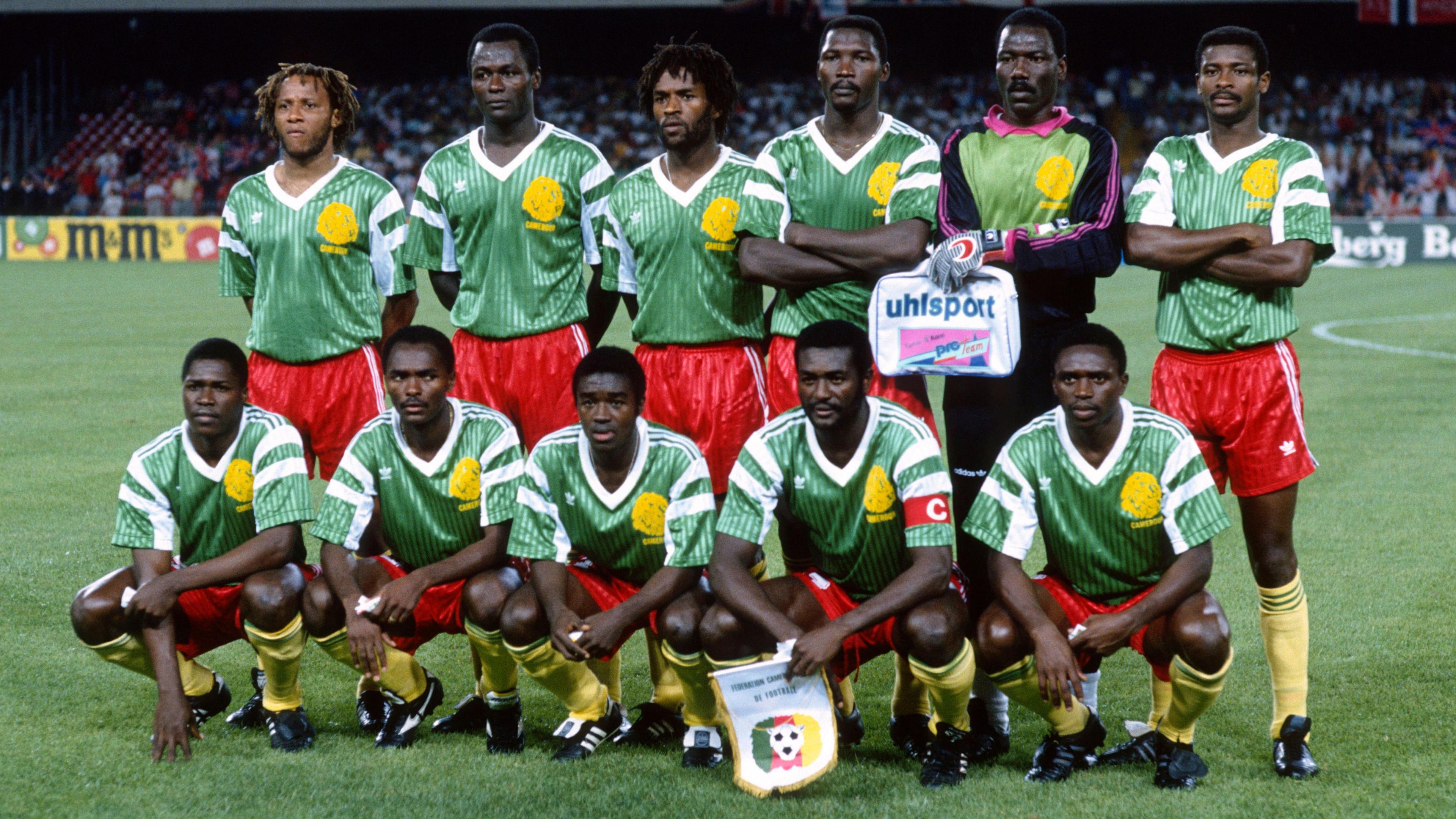 The Italy 1990 Cameroon World Cup team poses for a pre-match photograph on a green pitch. Six players stand in the rear, either with arms folded or hands clasped behind their back, including the goalkeeper second from the right. Five more players squat on their haunches in front, including the captain, also second from right, who wears a red captain's armband on his right upper arm and clutches a team pennant. The 10 outfield players all wear yellow socks, red shorts and green shirts with white trim and a large yellow lion on the left breast. The goalkeeper wears black tracksuit bottoms and a shirt which features black sleeves with vivid pink lines and a green chest which also includes the lion. He already has on his goalkeeping gloves and is clutching a small branded kit bag